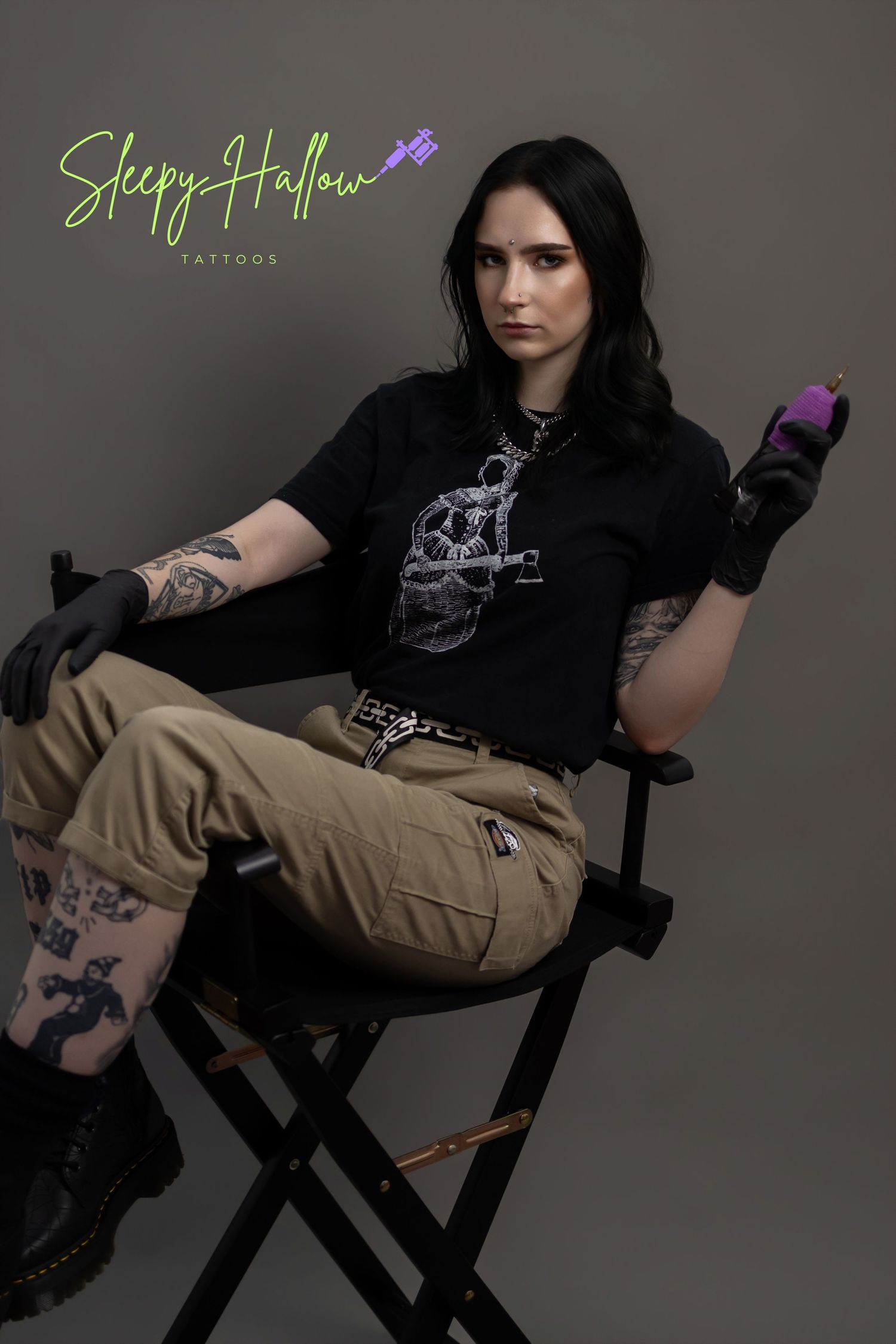 Tattooed tattoo artist sits in director's chair holding purple tattoo machine, wearing black graphic tee and cargo pants.