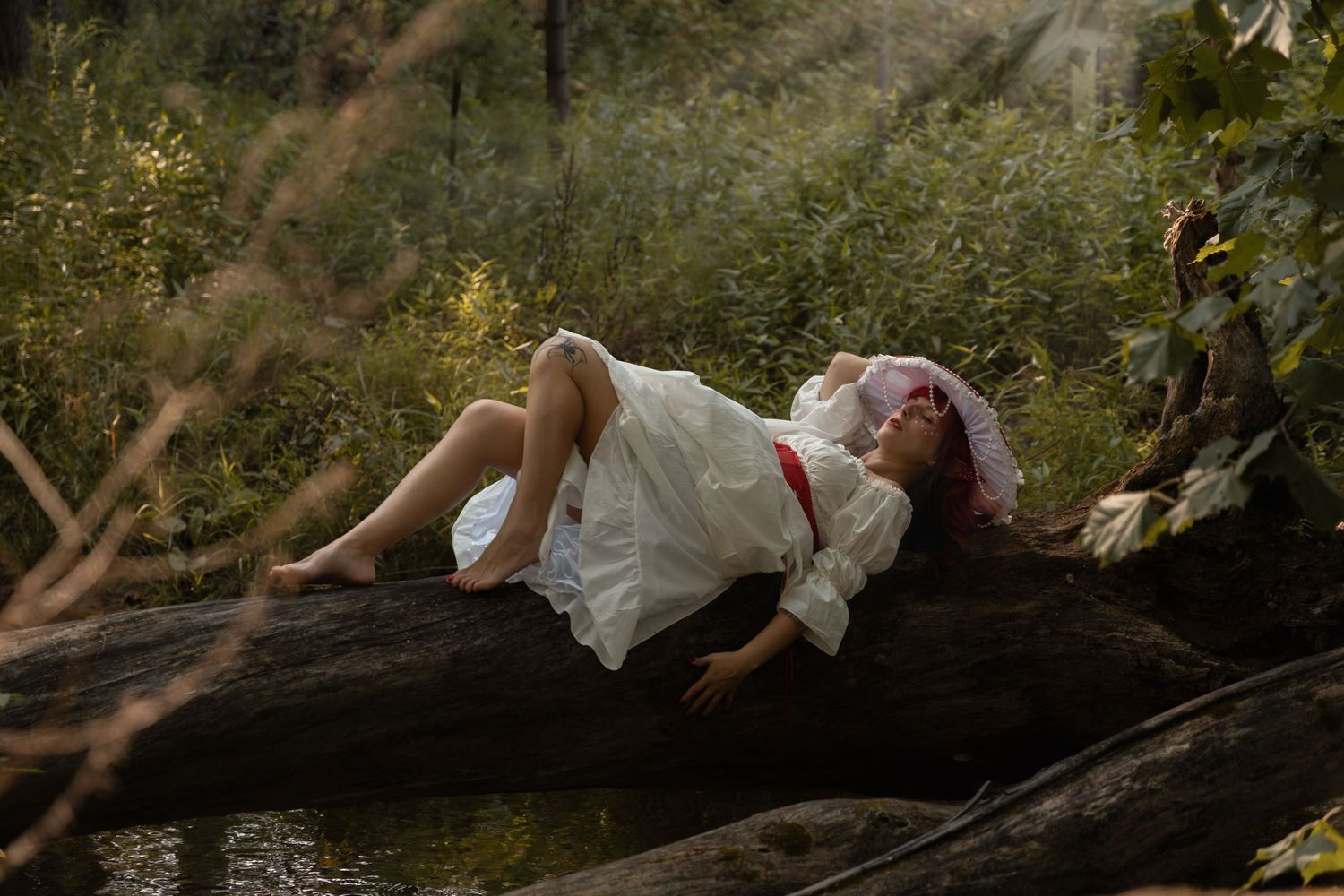 A barefoot girl in a white dress and red floral hat rests on a fallen log surrounded by lush green forest.