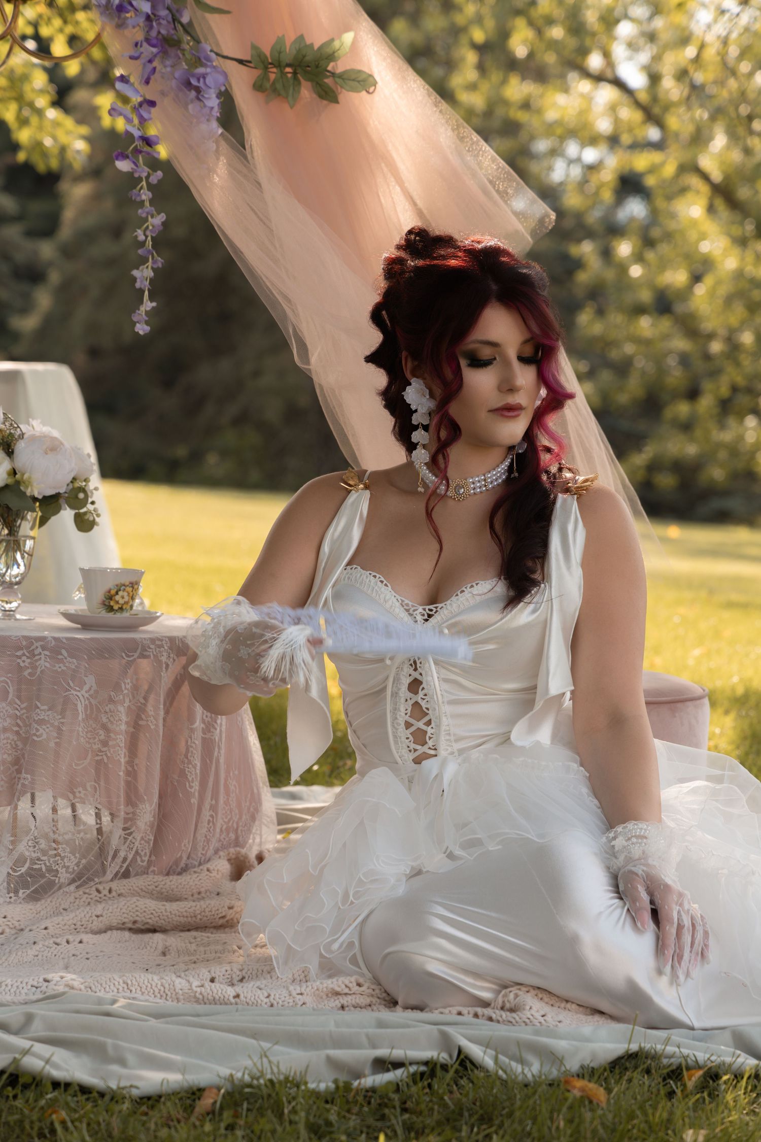 A bride in a white corset gown and gloves sits gracefully on a blanket outdoors near a tea table with wisteria decor.