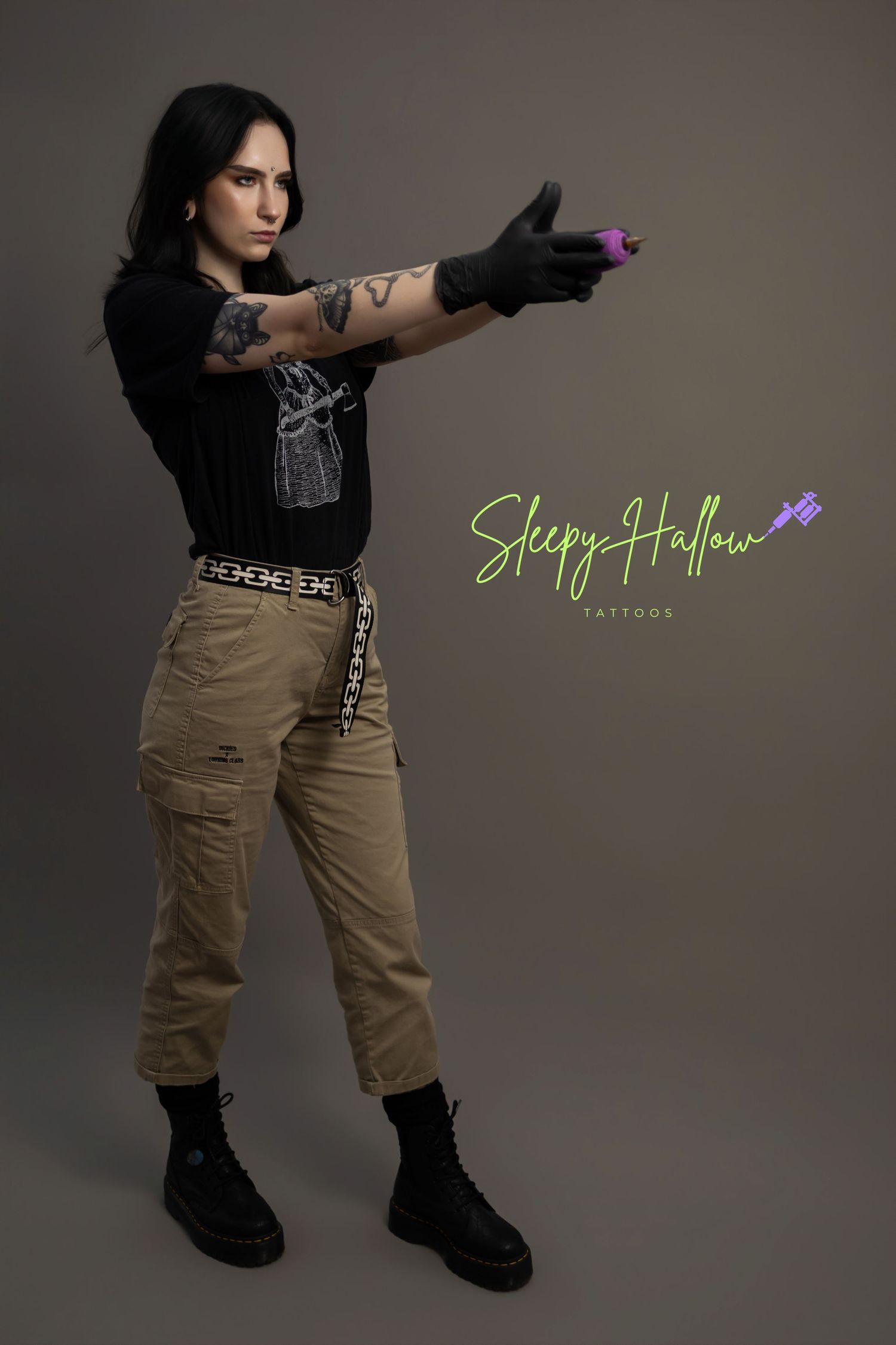 Tattooed woman in black tee and cargo pants aims a purple tattoo gun, promoting Sleepy Hallow Tattoos.