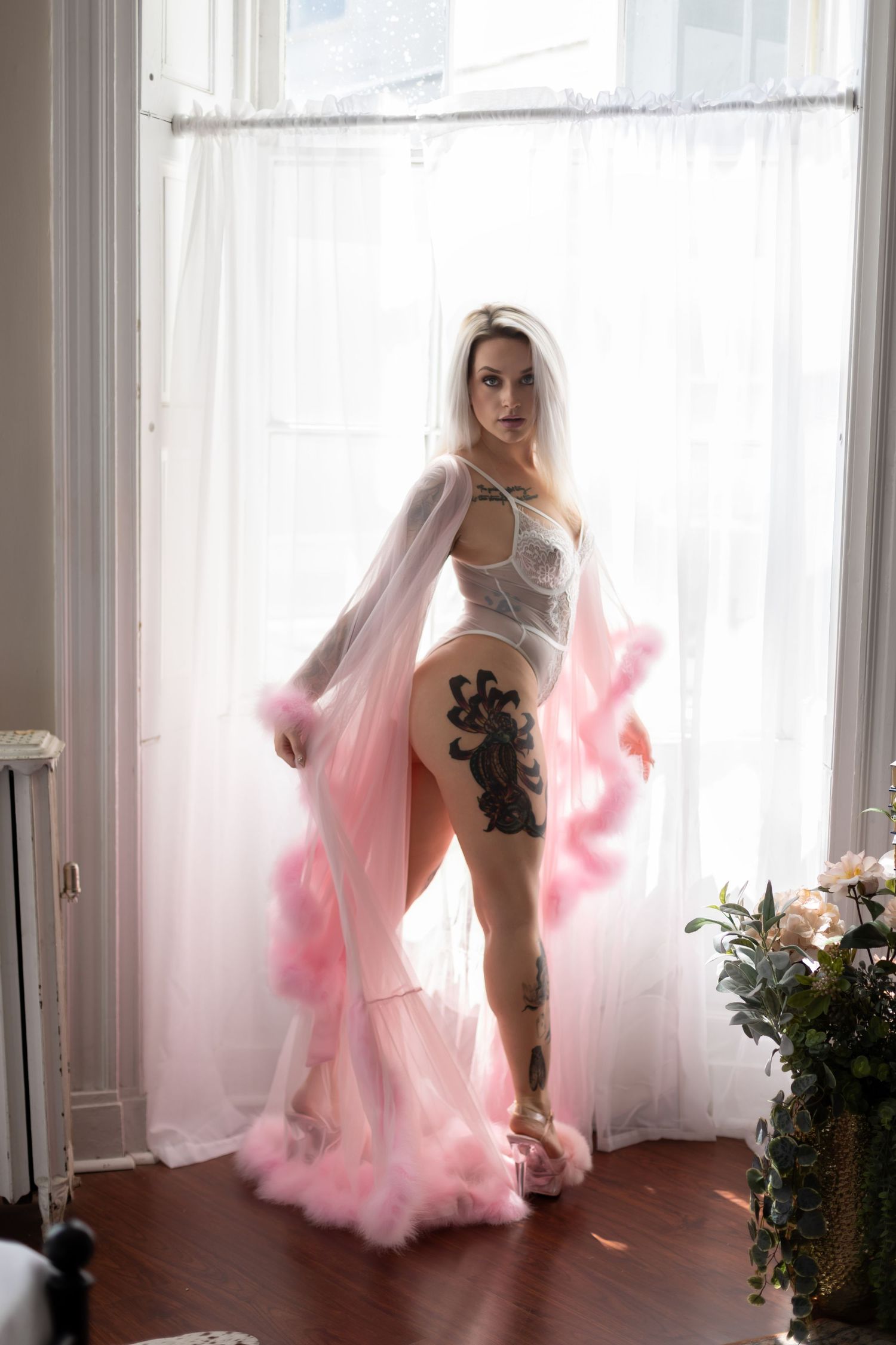 Blonde woman with tattoos wearing white lingerie and pink feather robe posing by a bright window.