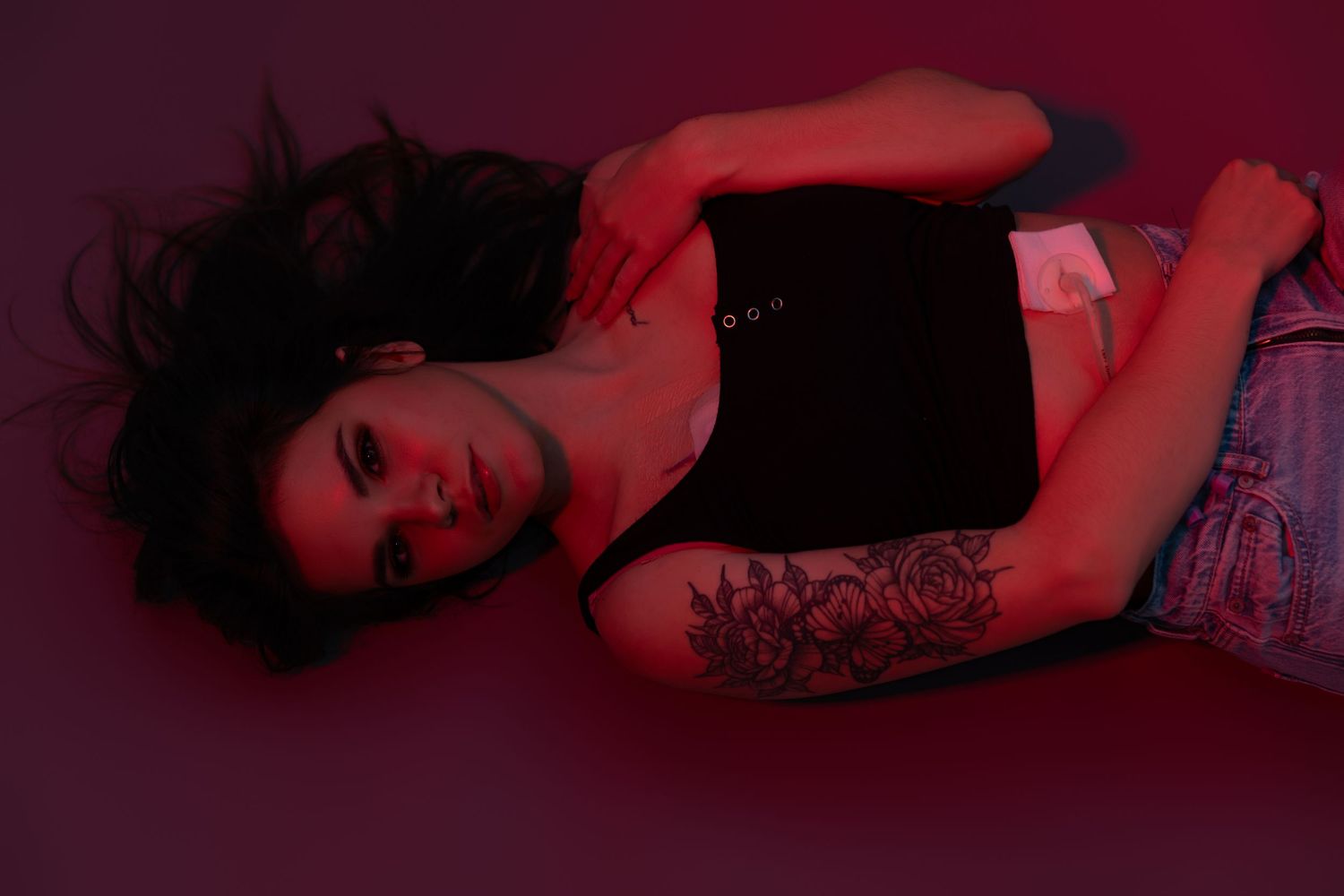 Woman with tattoos lying on floor under red lighting, wearing black tank top and denim shorts.