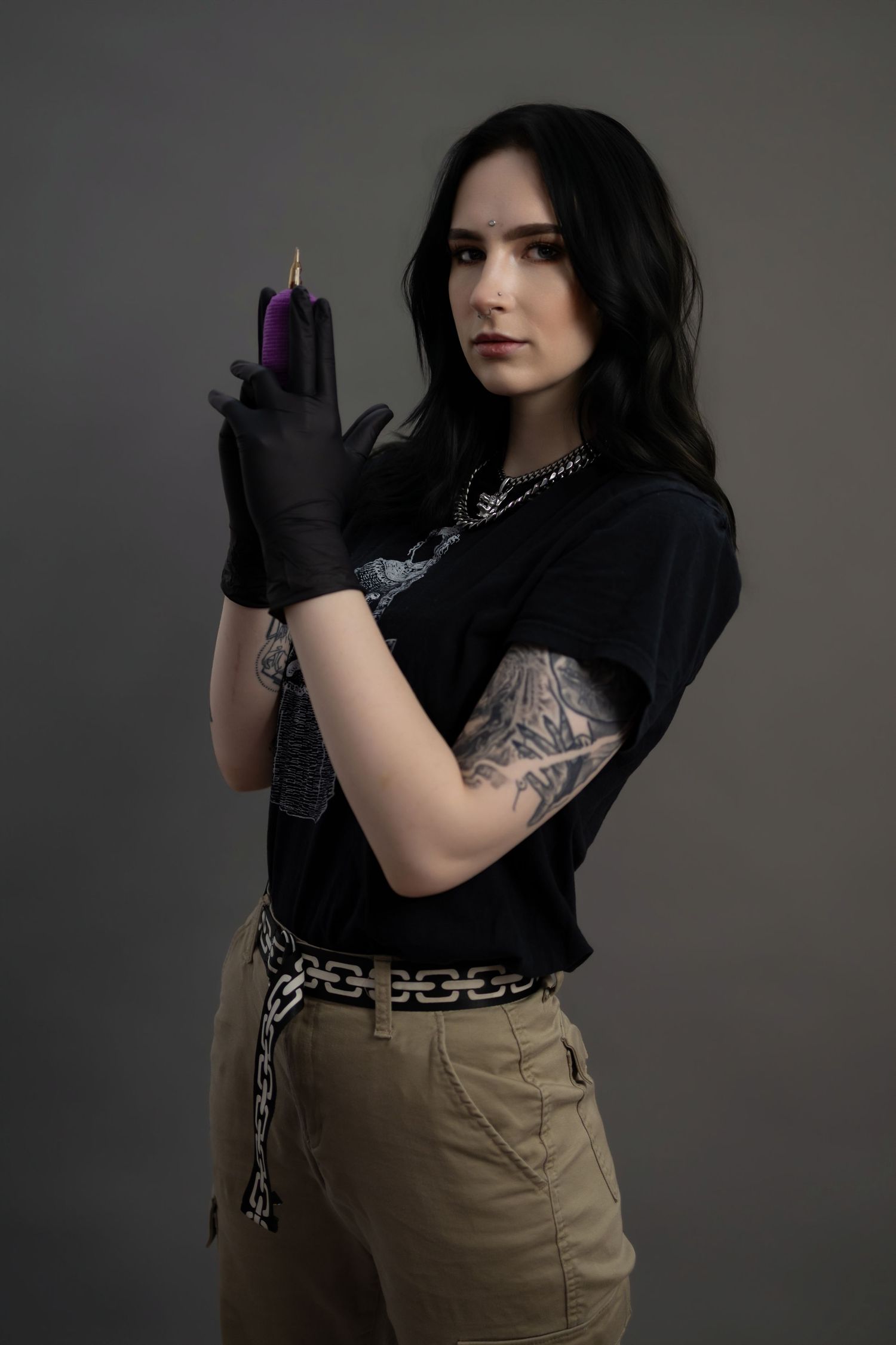 Tattooed woman in black gloves holds a purple tattoo machine in a professional studio portrait.