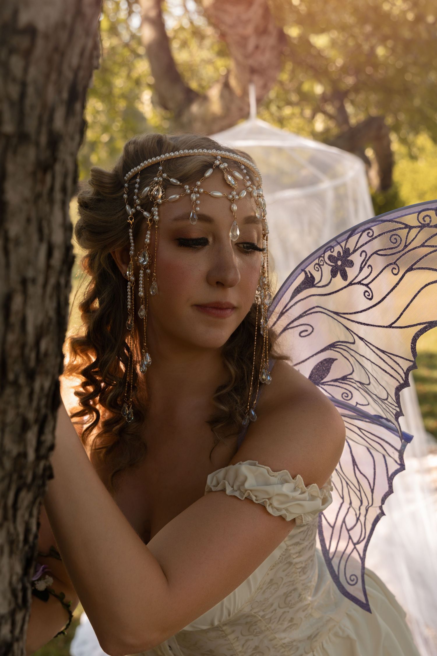 A woman in a white off-shoulder gown with fairy wings and pearl headpiece poses dreamily in a sunlit forest.