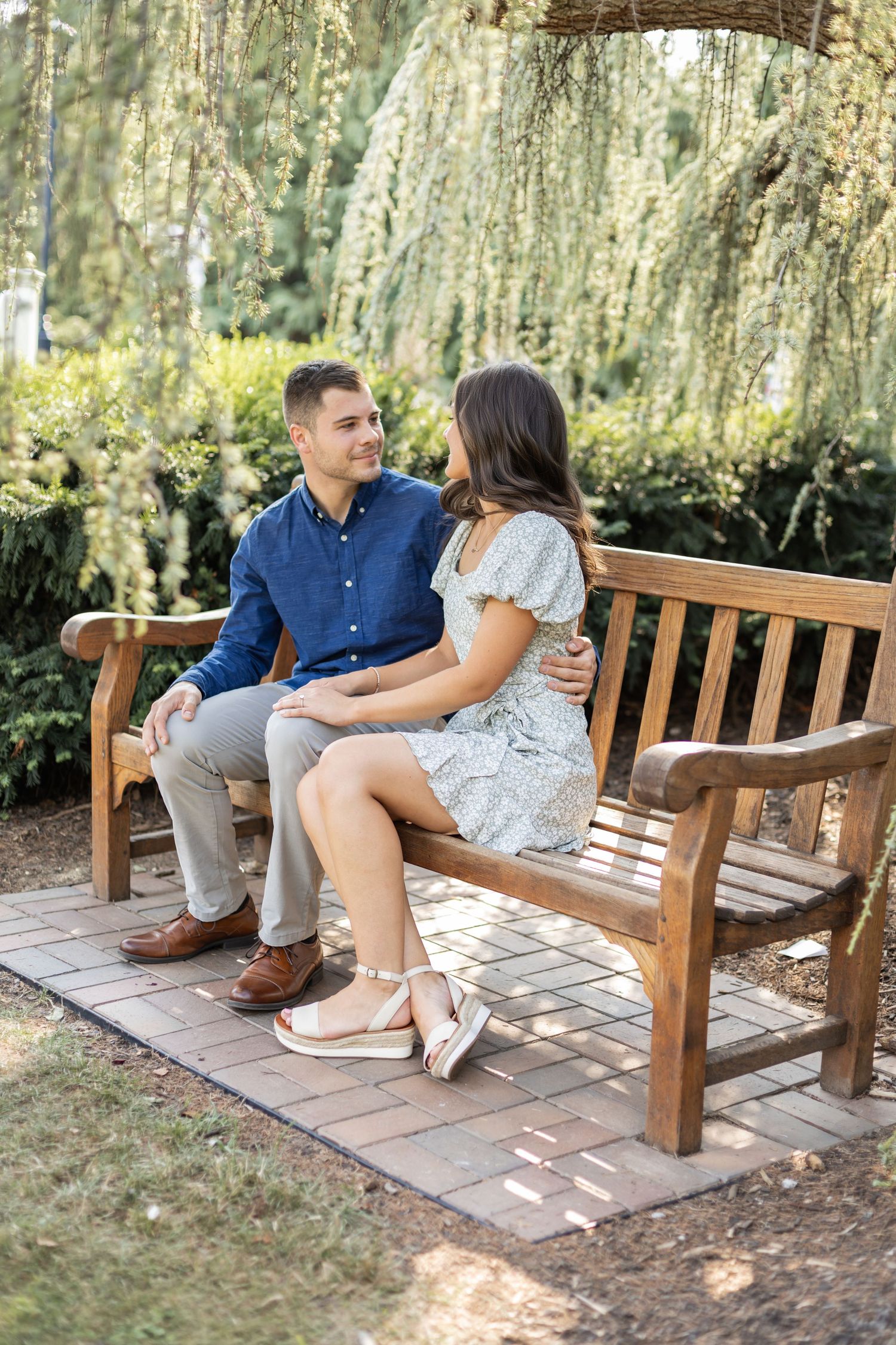 A couple shares a romantic moment while sitting on a wooden bench under a weeping willow tree in a garden setting.