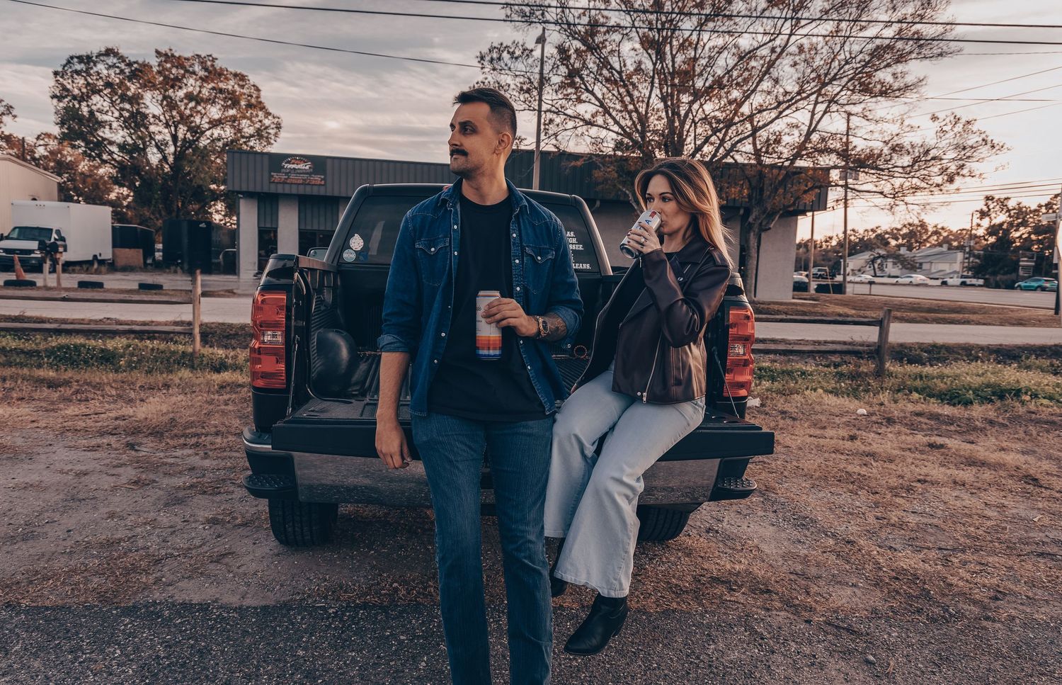 Two people lean against a pickup truck at sunset in a casual, relaxed pose on dusty ground.