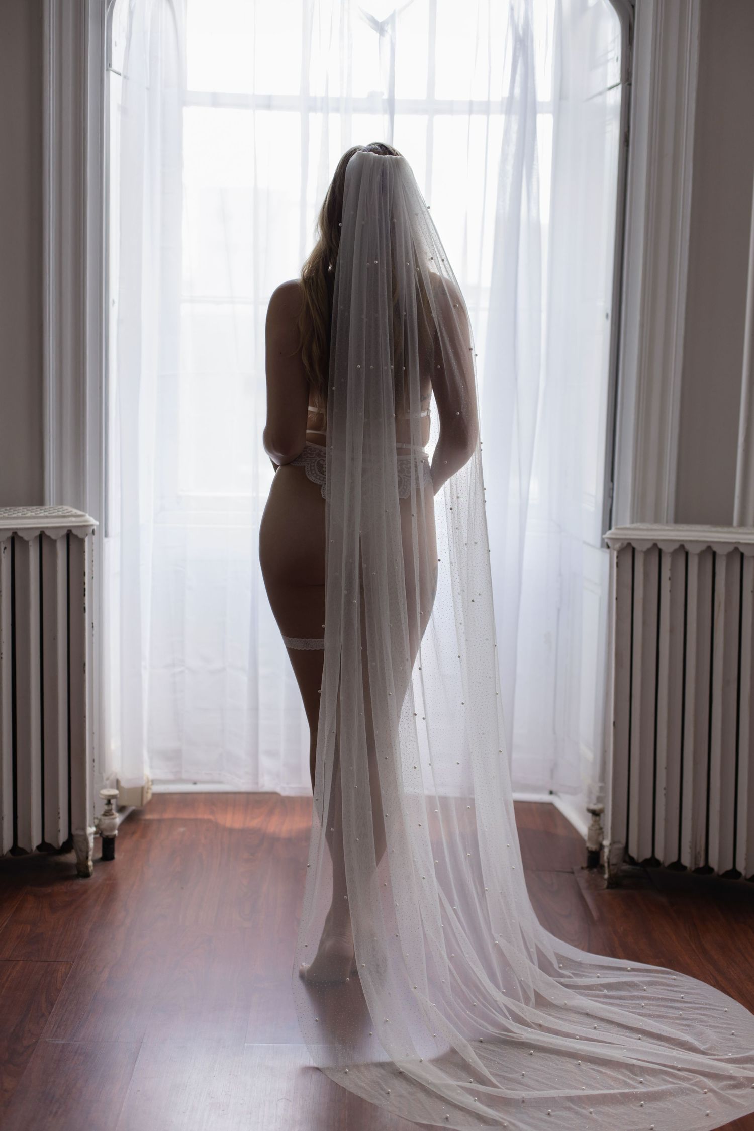 Bride in lingerie and long veil standing backlit by window light on hardwood floor.