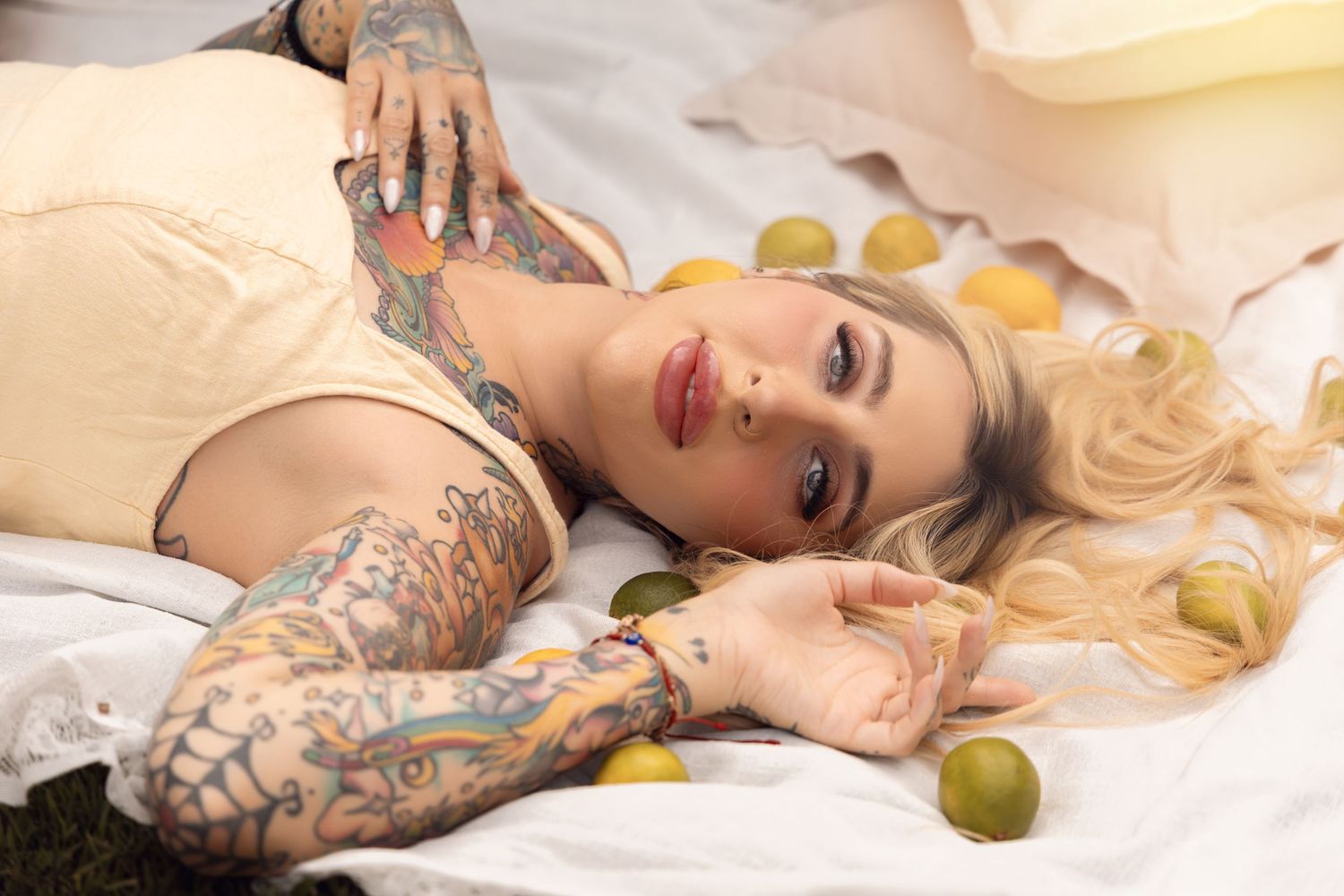 Tattooed blonde woman lying on white fabric surrounded by limes in a creative artistic portrait photo.