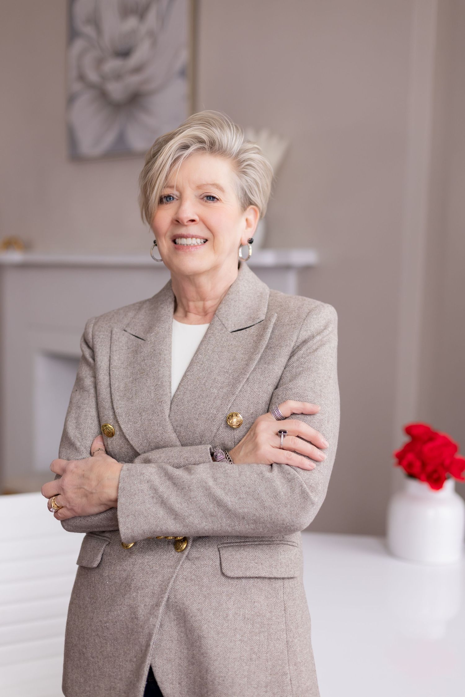 Confident woman in grey blazer poses with crossed arms in bright office space.