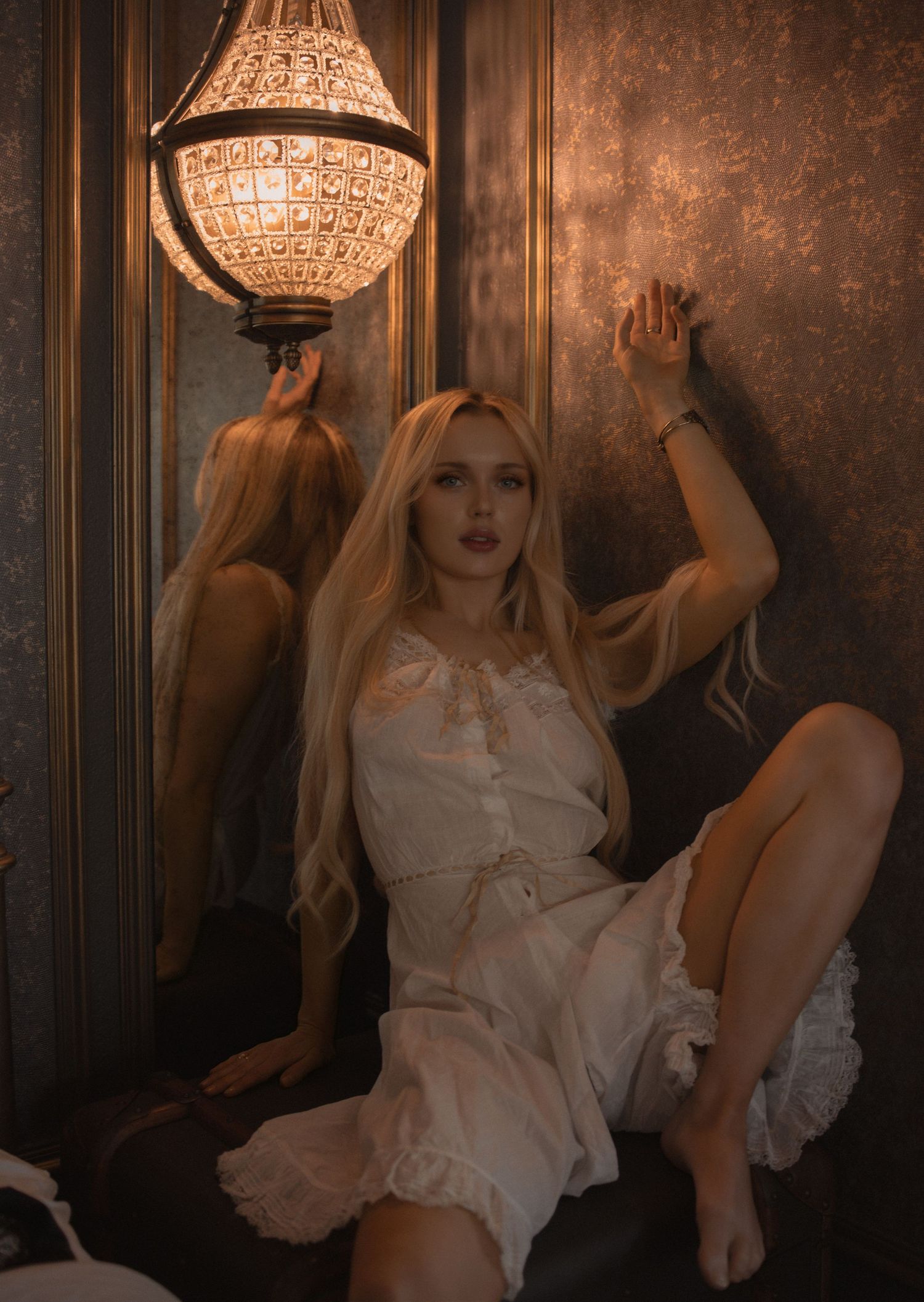 Blonde woman in white dress poses dramatically by ornate chandelier against dark golden wall.