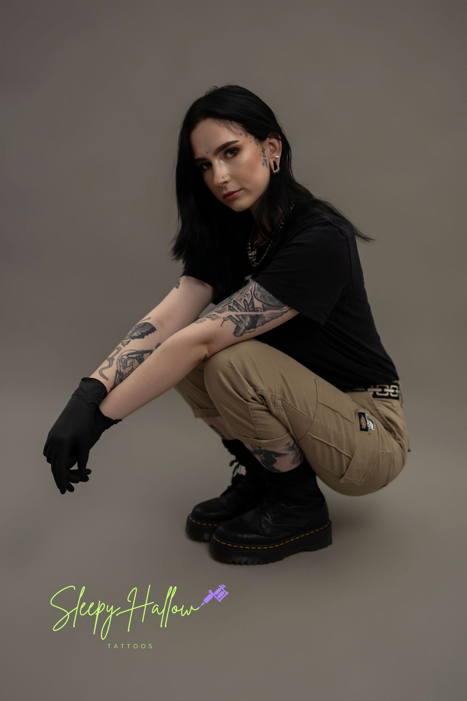 Tattooed woman crouching in black tee, khaki cargo pants, and black boots with gloves, studio photo.