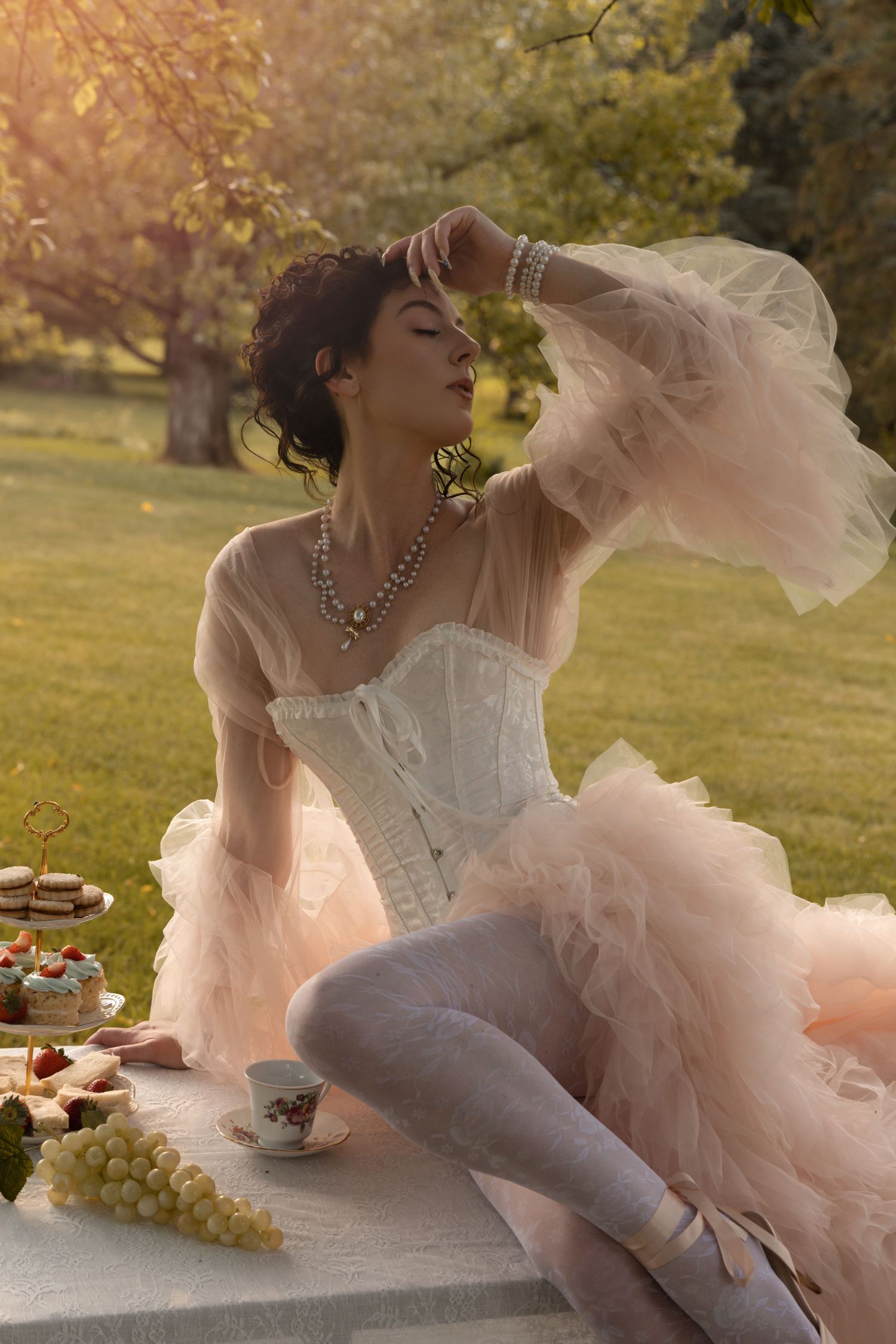 Woman in white corset and pink tulle skirt at outdoor tea party with grapes and pastries in golden light.