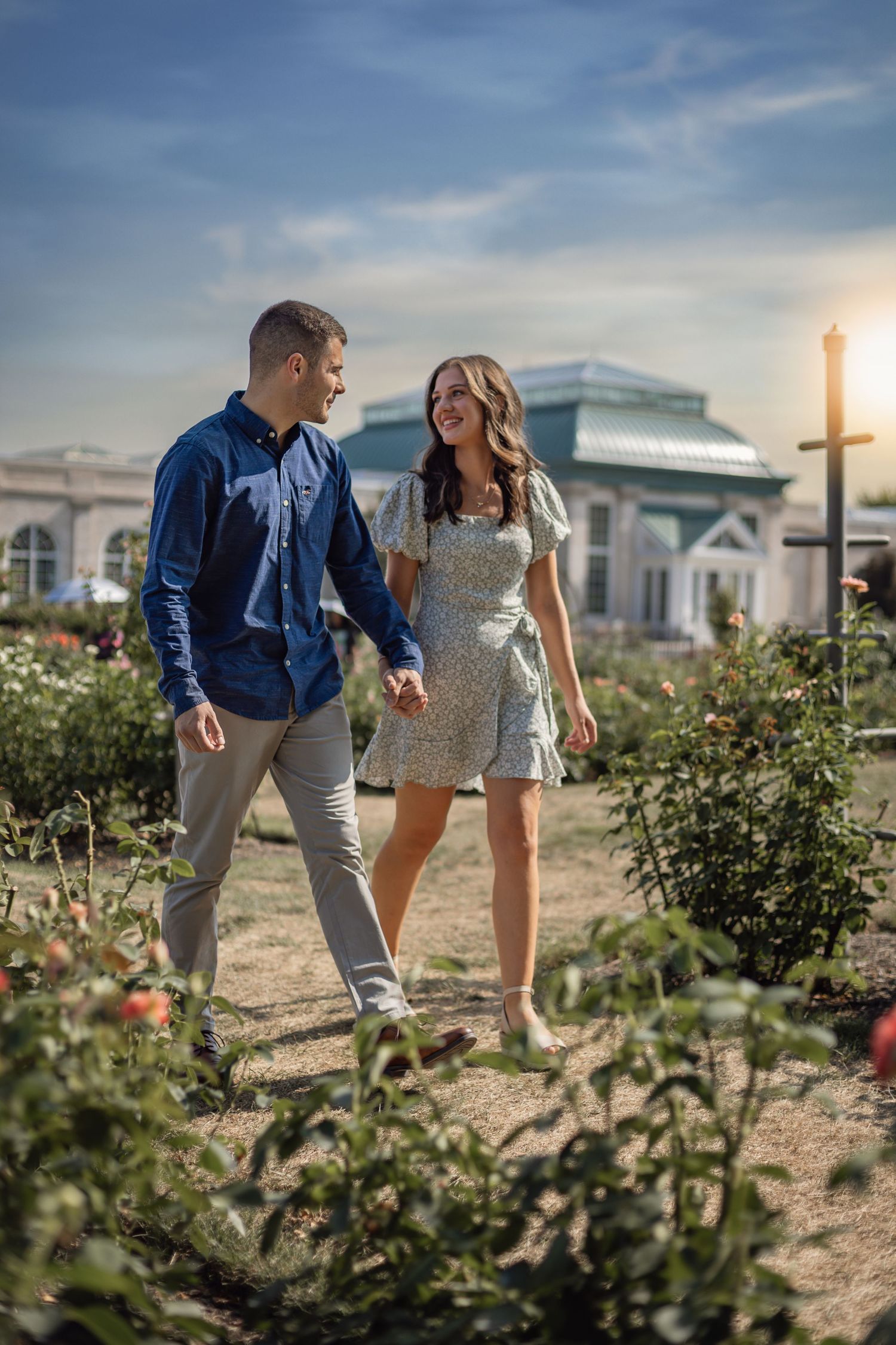 A couple walks hand in hand through a garden path with a white building in the background at sunset.