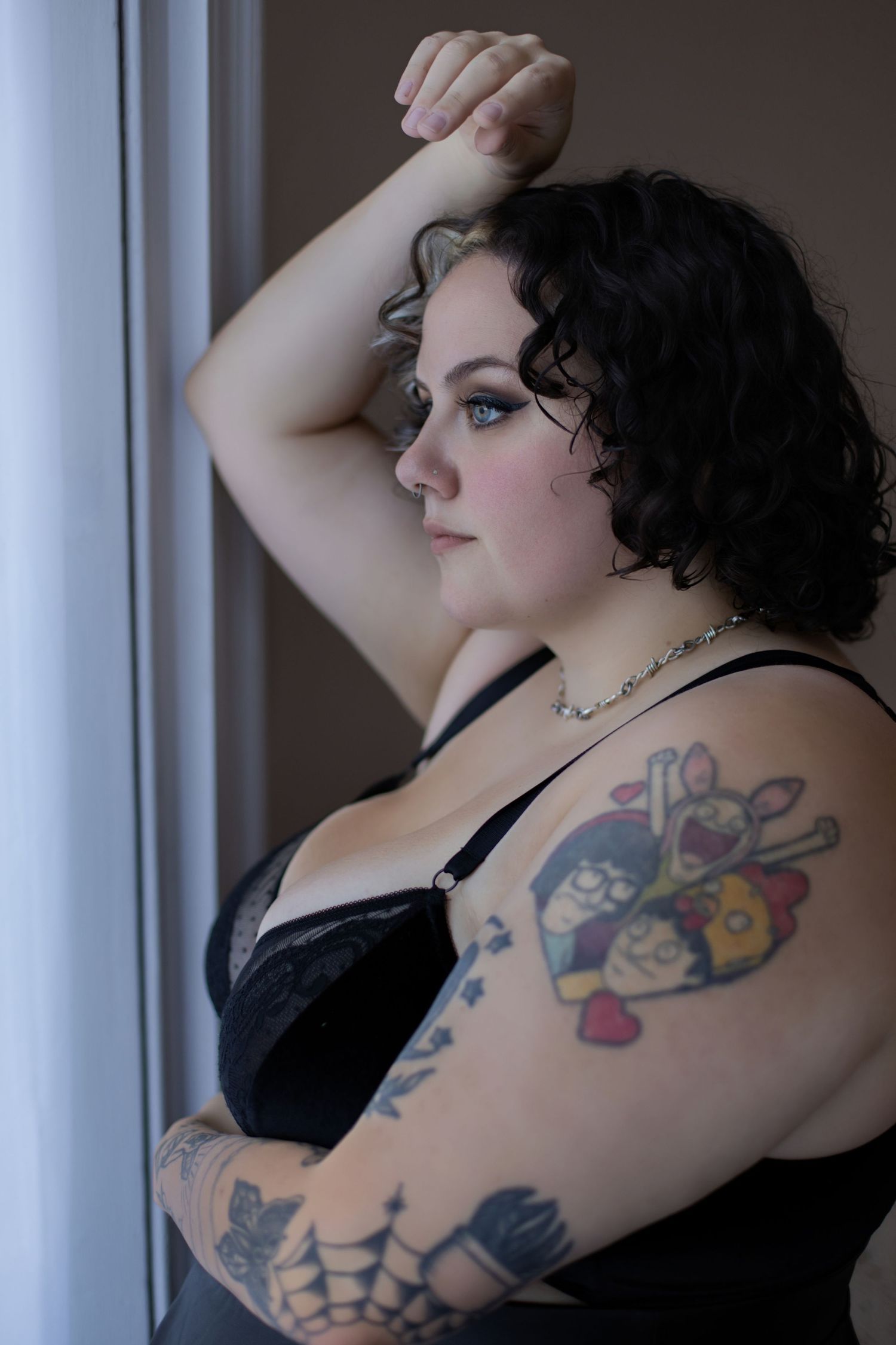 A curly-haired figure in black lingerie poses by a window, showing colorful tattoos on their arm.