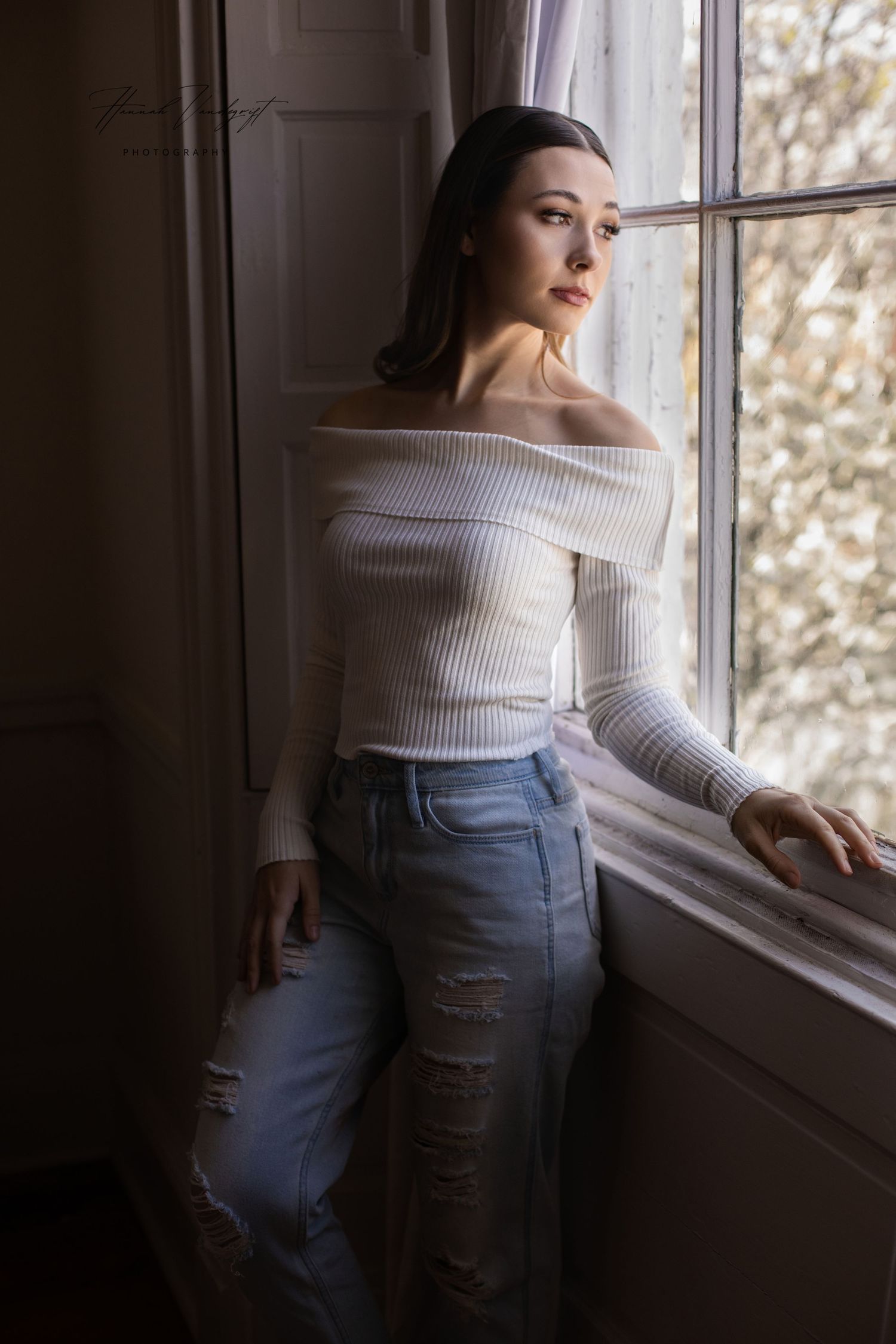Figure in white sweater and blue jeans leans thoughtfully against window during portrait session.