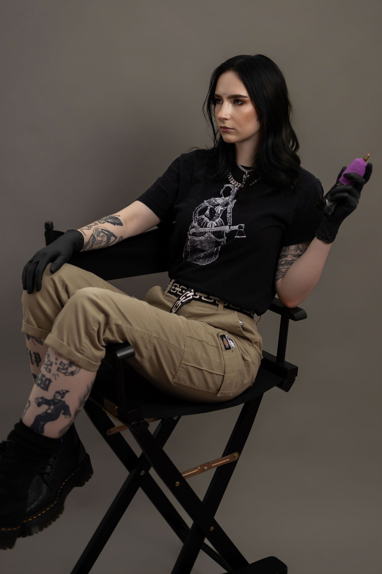 Tattooed woman in black graphic tee and cargo pants sits in director's chair holding purple object, wearing black gloves.