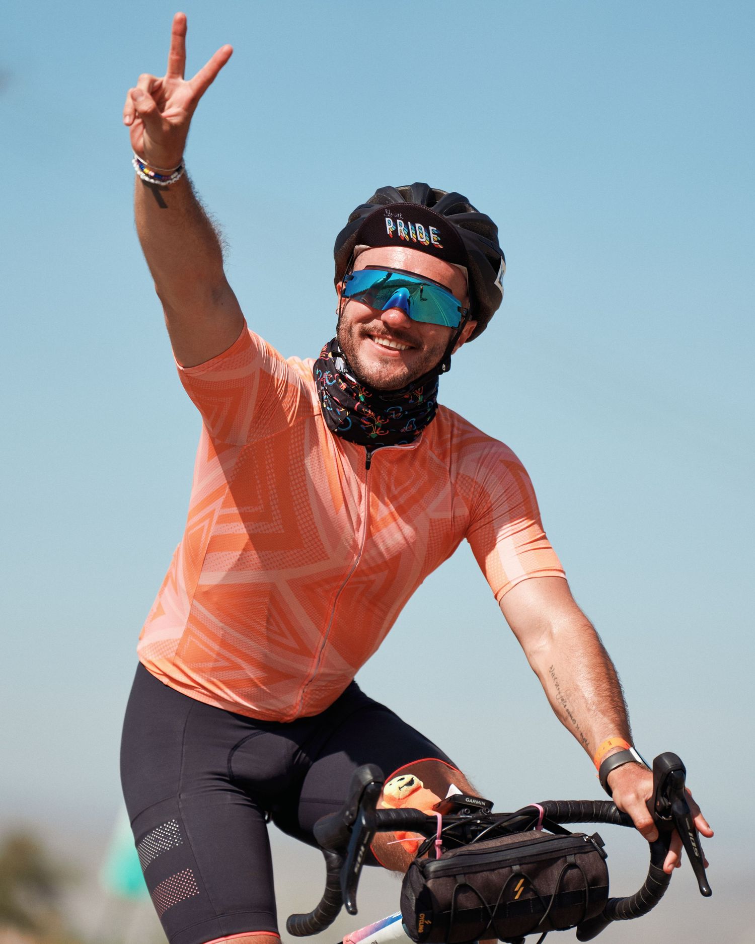 Cyclist in peach-colored jersey makes peace sign while riding bike against blue sky.