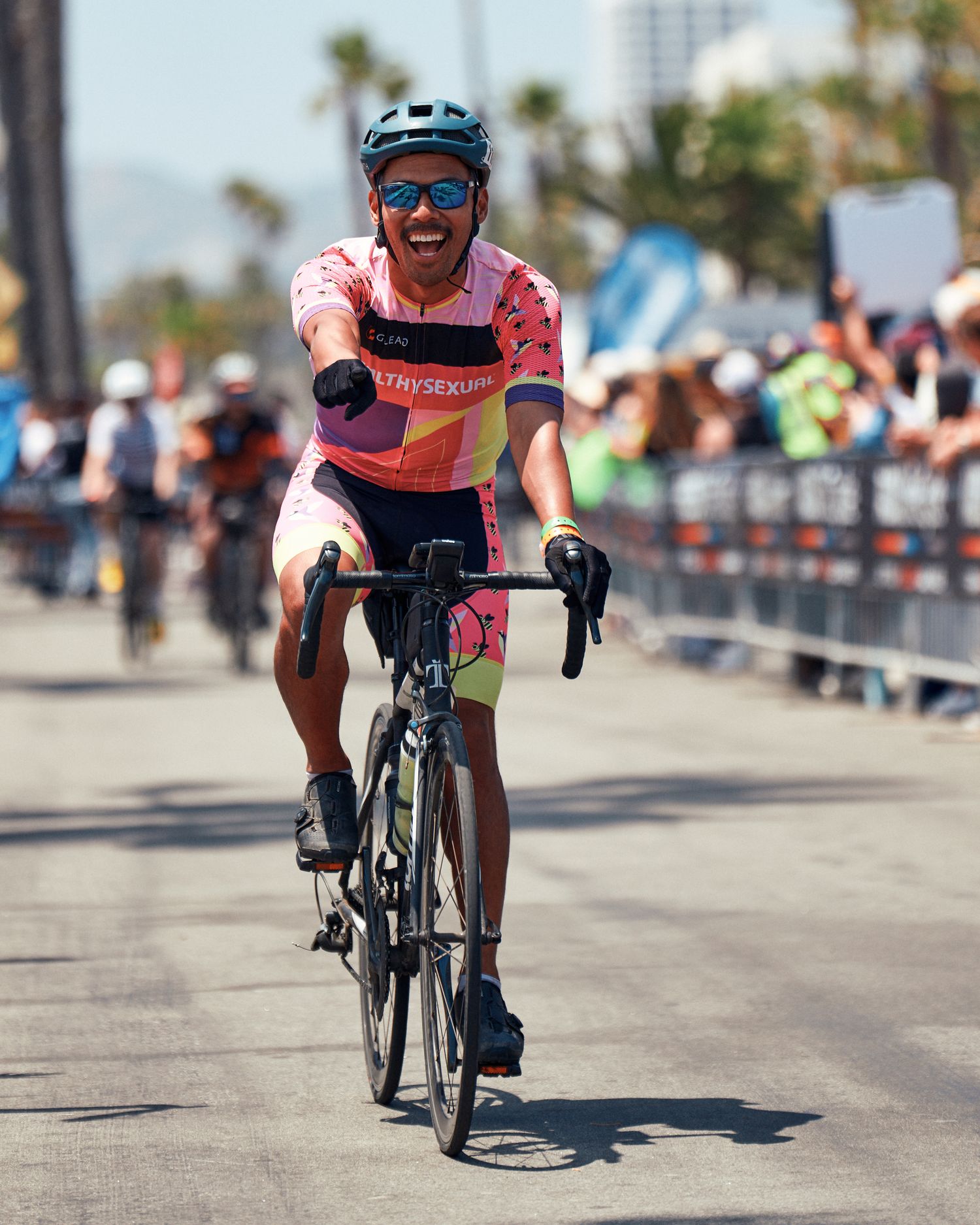 A cyclist in racing gear rides a black road bike on a paved course with palm trees visible in the background.
