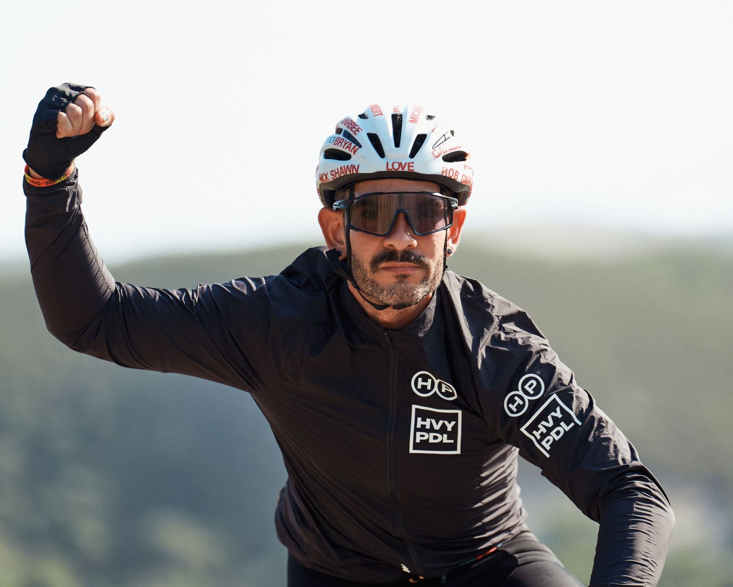 A cyclist in black cycling gear with sunglasses and helmet raises their arm while riding outdoors against a blurred background.