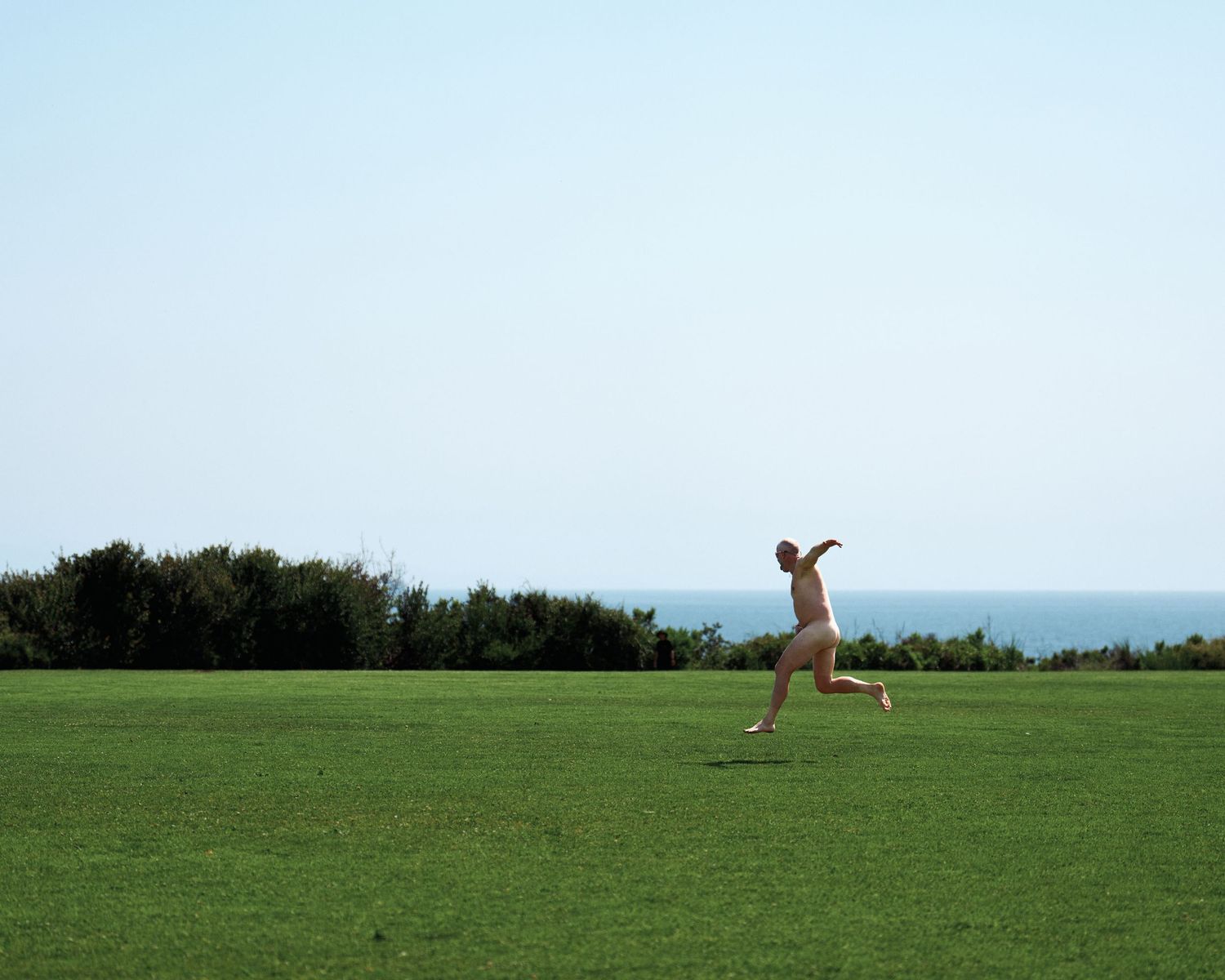 Person running joyfully across a green hilltop field against blue sky and ocean vista.