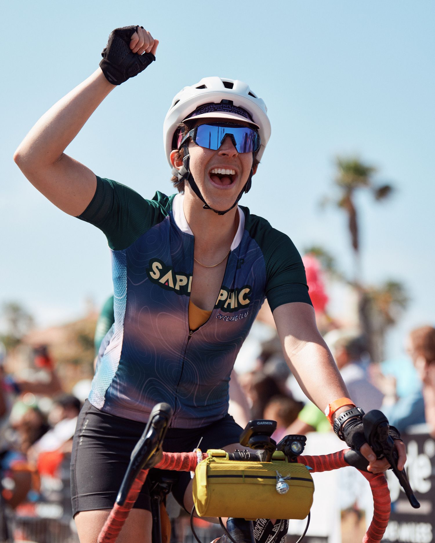 Cyclist in a navy cycling jersey and helmet celebrates victory with raised fist against blue sky and palm trees.