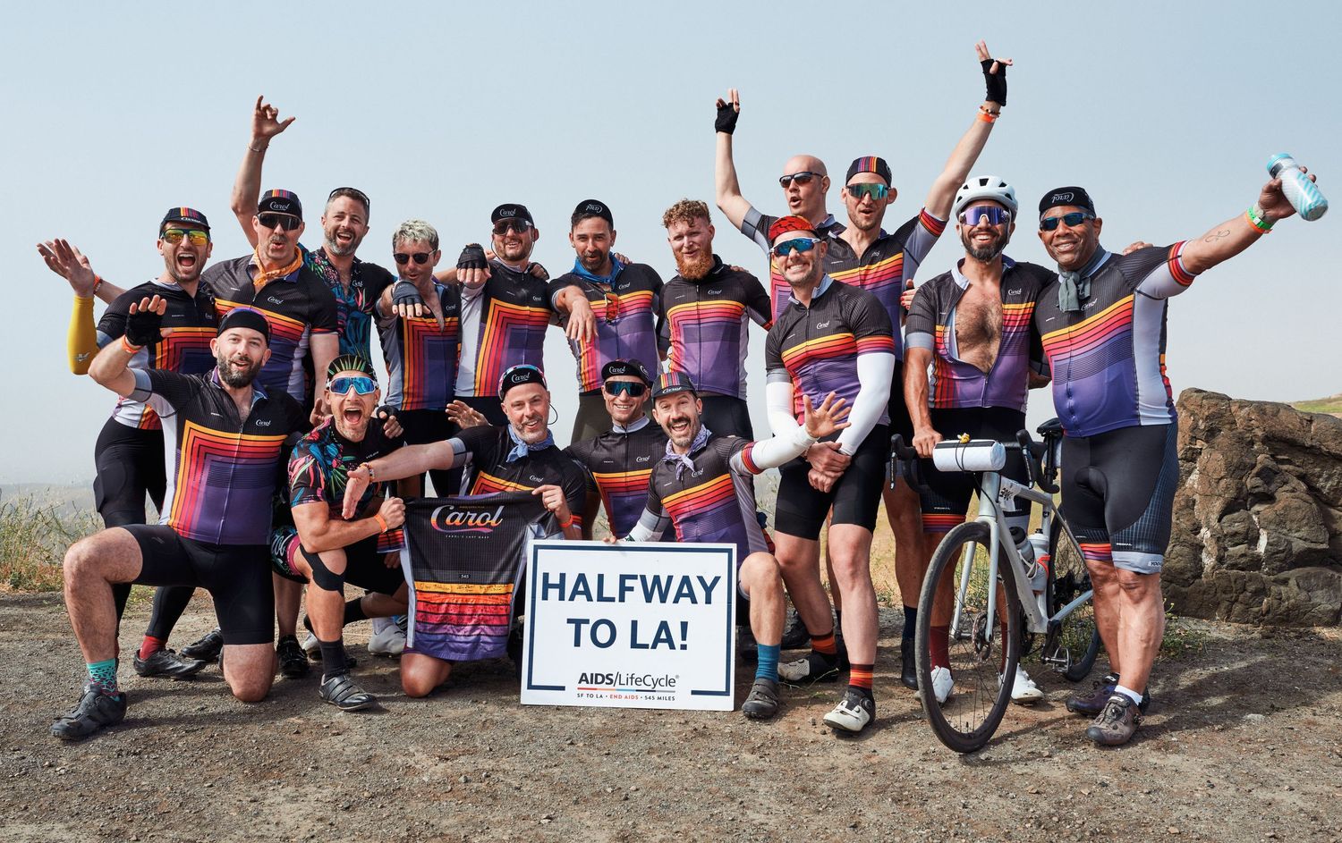 Group of cyclists in matching jerseys pose with Halfway to LA sign during AIDS LifeCycle ride.