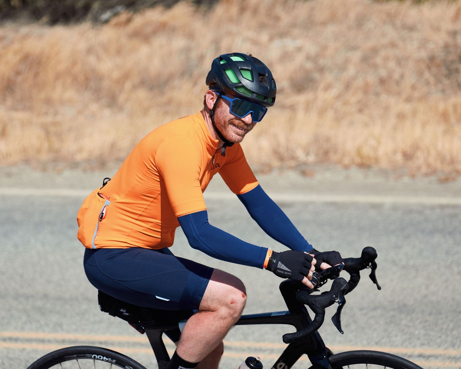 Cyclist in bright orange jersey and navy shorts rides road bike on scenic mountain road.
