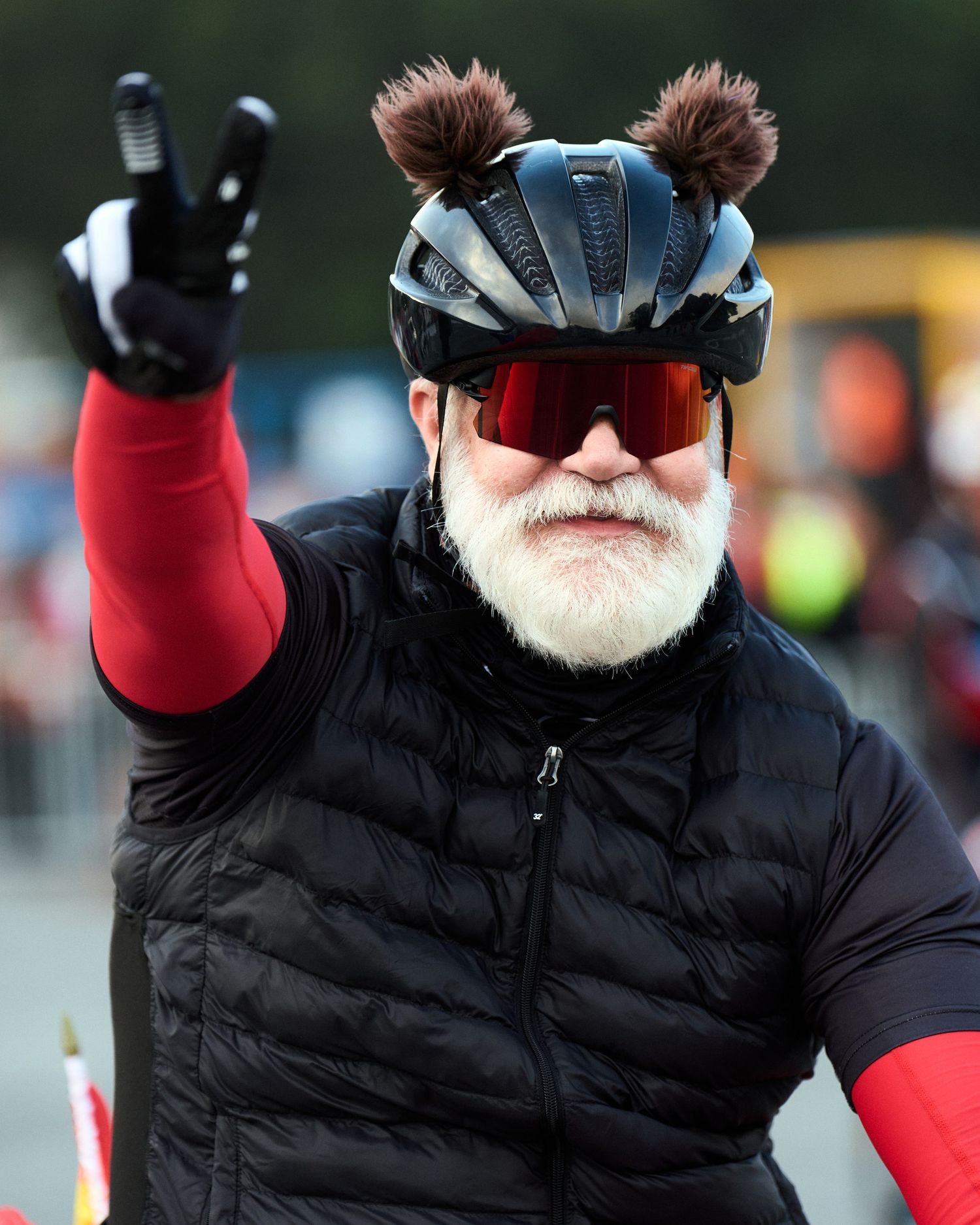 Cyclist in winter gear with furry ear attachments on helmet giving peace sign during event.