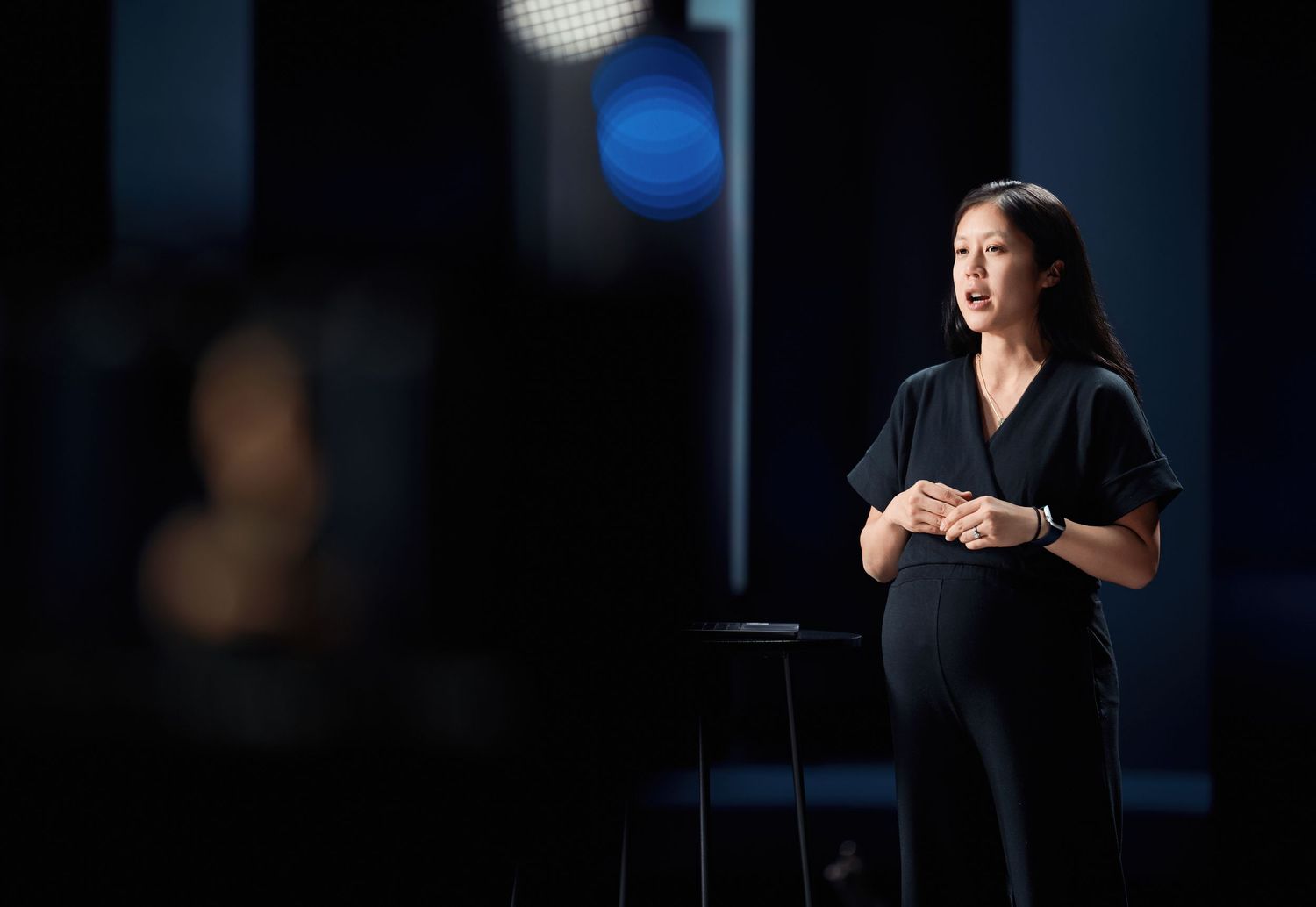 Against a dark background with blue lighting, a speaker wearing black stands thoughtfully on a stage during a presentation.