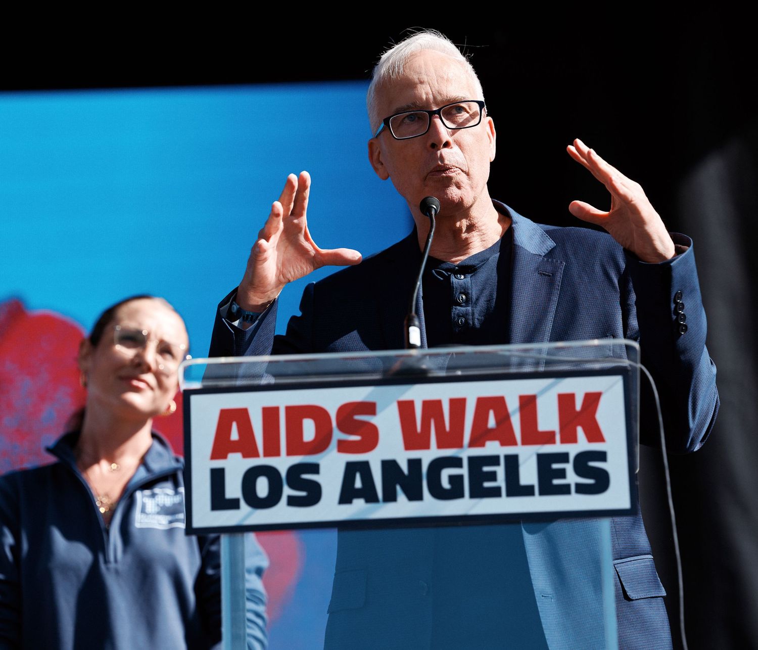 A person in glasses gestures while speaking at a podium during an AIDS Walk Los Angeles event.