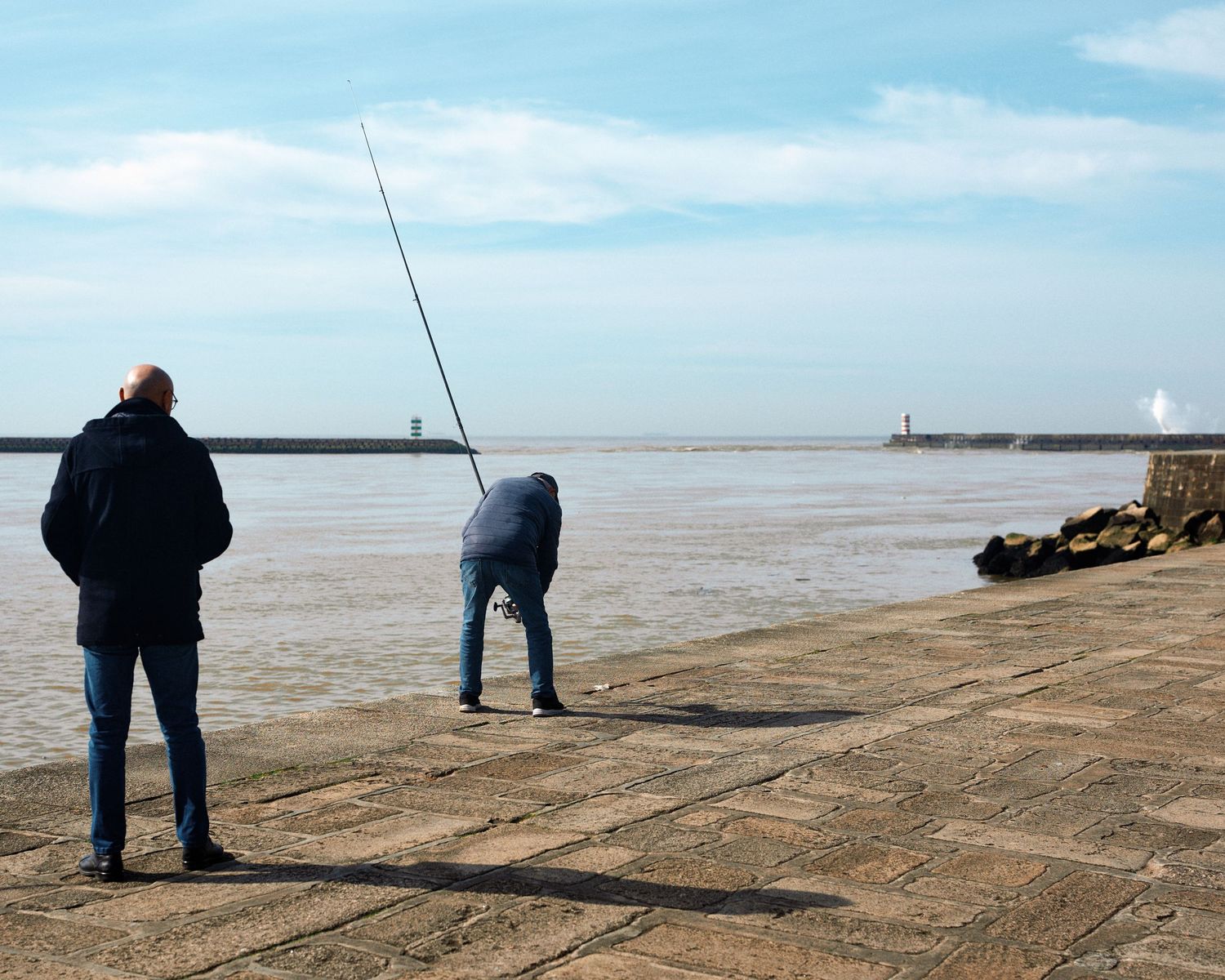 Fishermen stand along a concrete pier casting their lines into the ocean on a cloudy day.