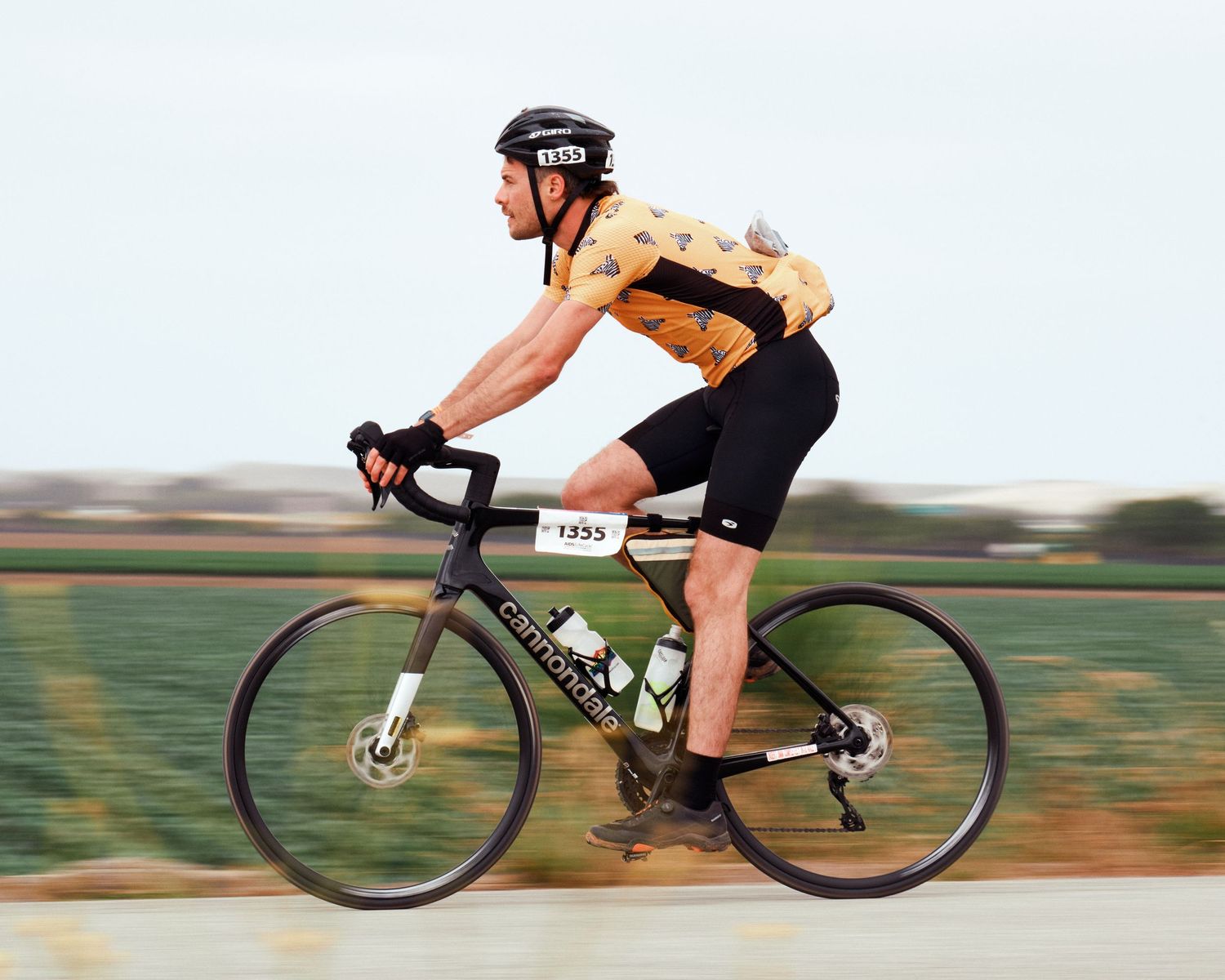 Cyclist in orange jersey riding carbon fiber road bike past blurred agricultural landscape.