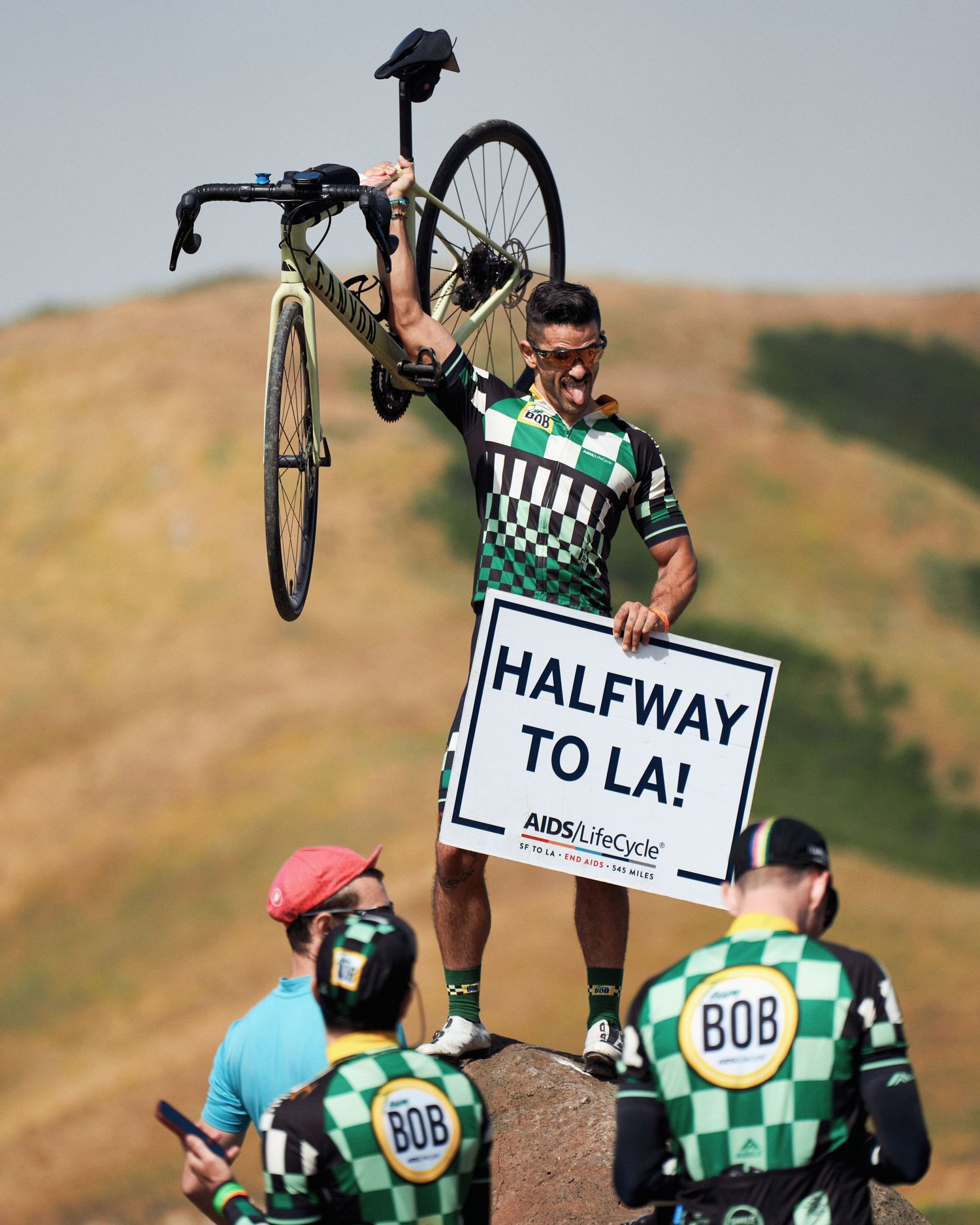 Cyclists in green checkered jerseys holding a 'Halfway to LA' sign on a hilltop during a group ride.