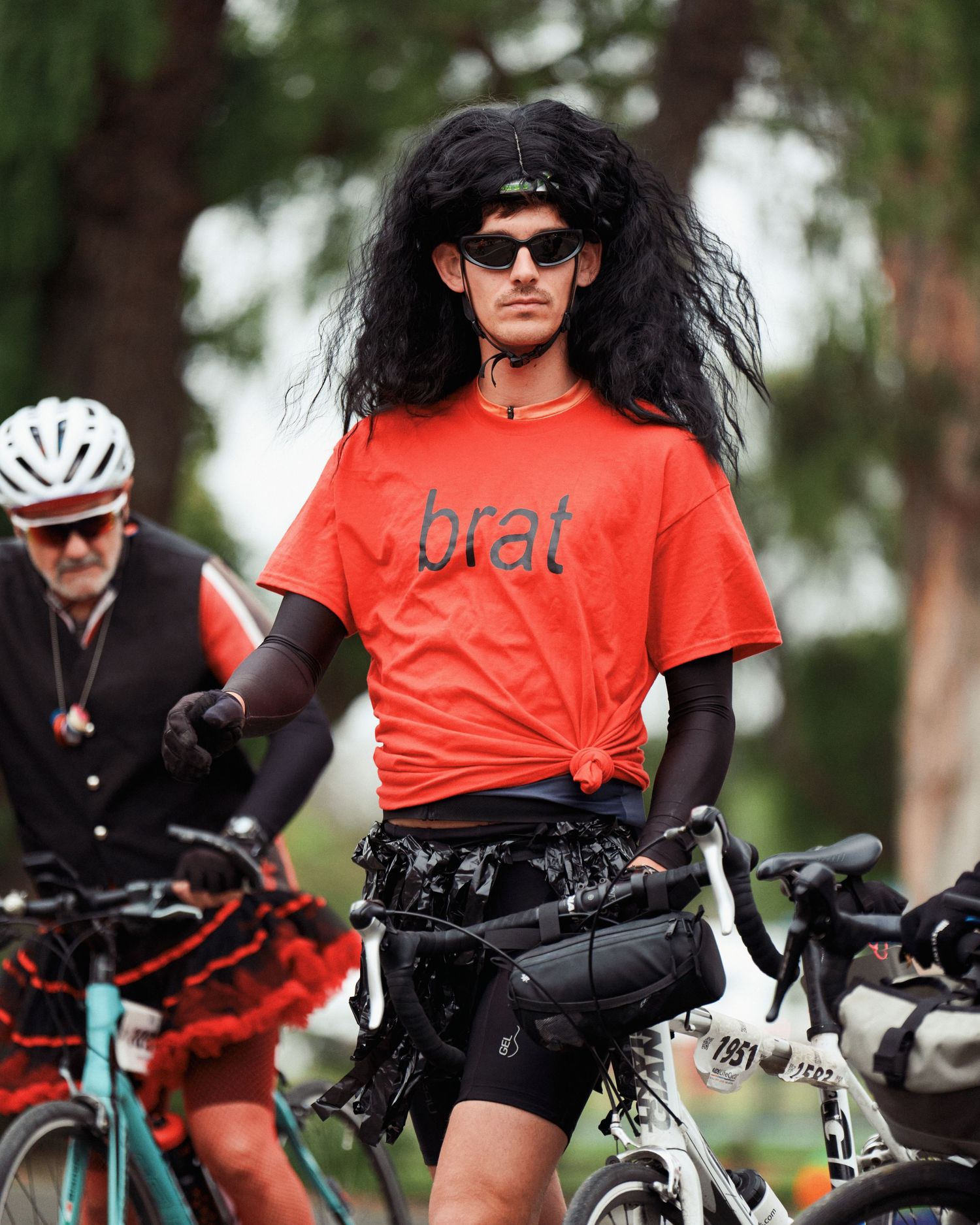 Cyclist wearing orange 'brat' t-shirt and long black wig poses with bicycle.