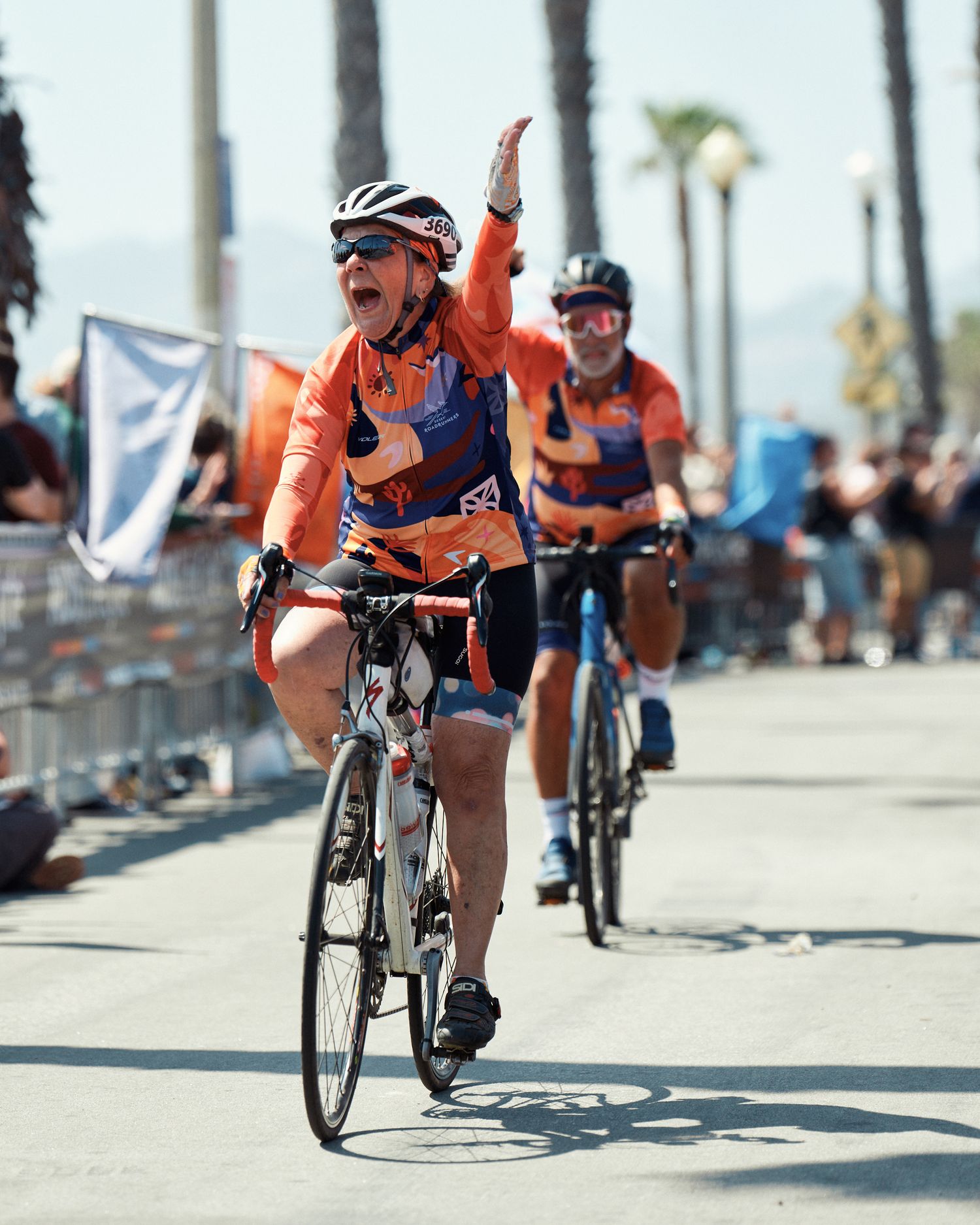 Cyclists in orange jerseys race along a palm-lined beachfront path during a competitive cycling event.