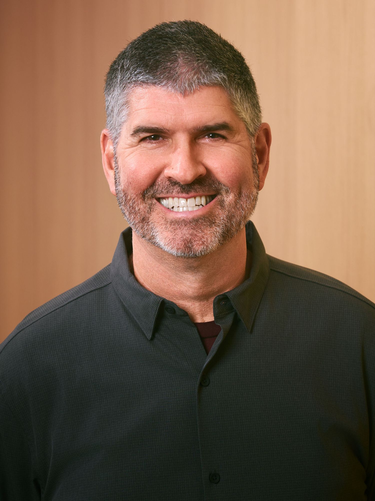 Smiling middle-aged man with short gray hair and beard wearing a black polo shirt against a warm wooden background.