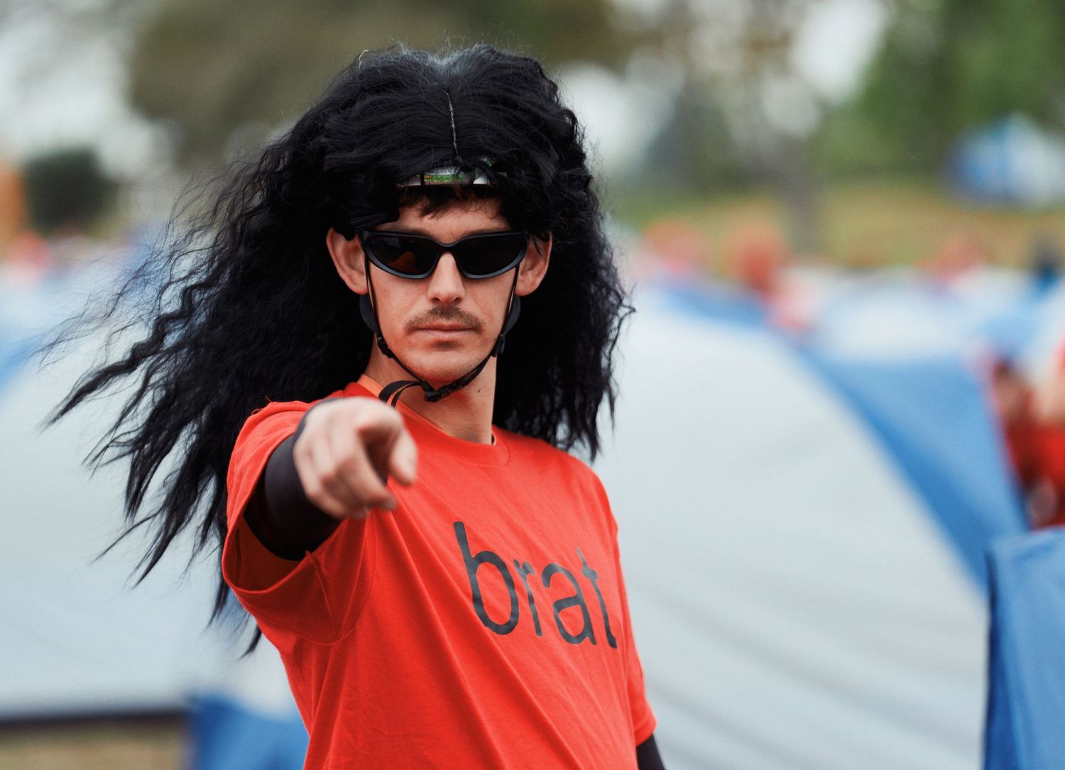 A performer with dark hair and sunglasses wearing a red t-shirt jumps energetically at an outdoor festival event.