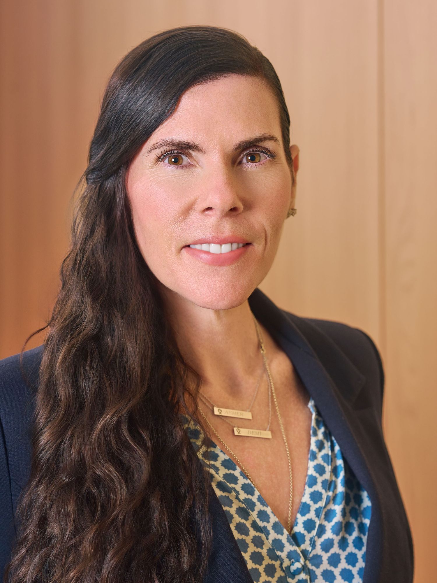 Professional headshot of a woman with long dark hair wearing a blue blazer and layered gold necklaces.