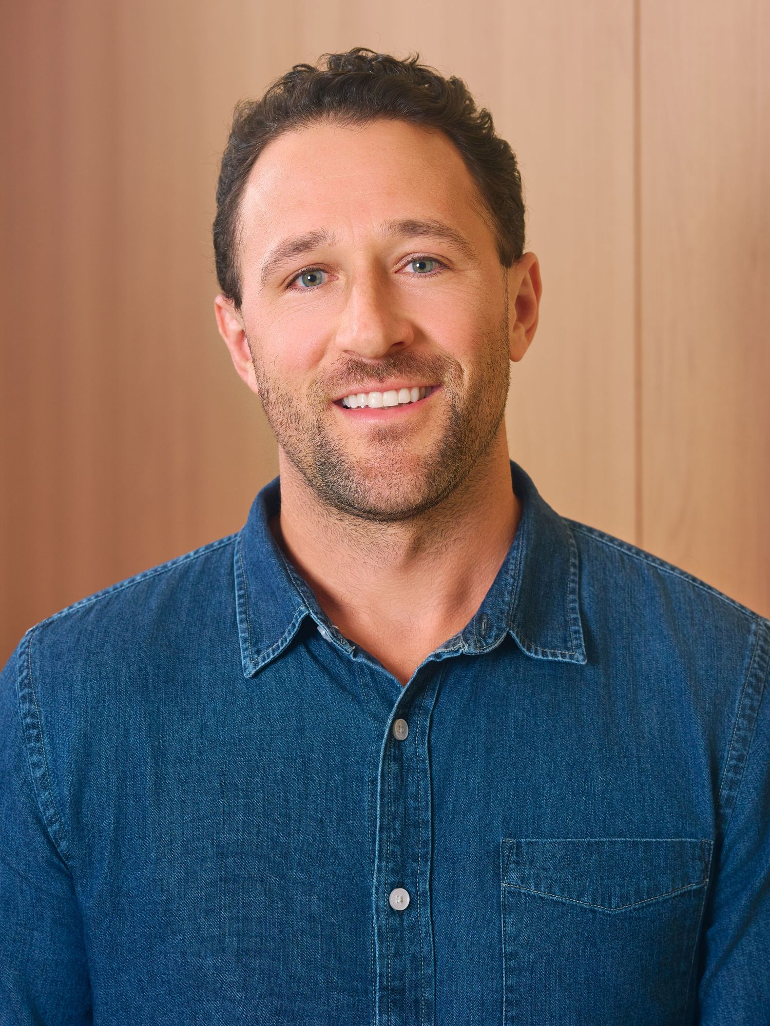 A person wearing a denim button-up shirt smiles warmly against a beige background.