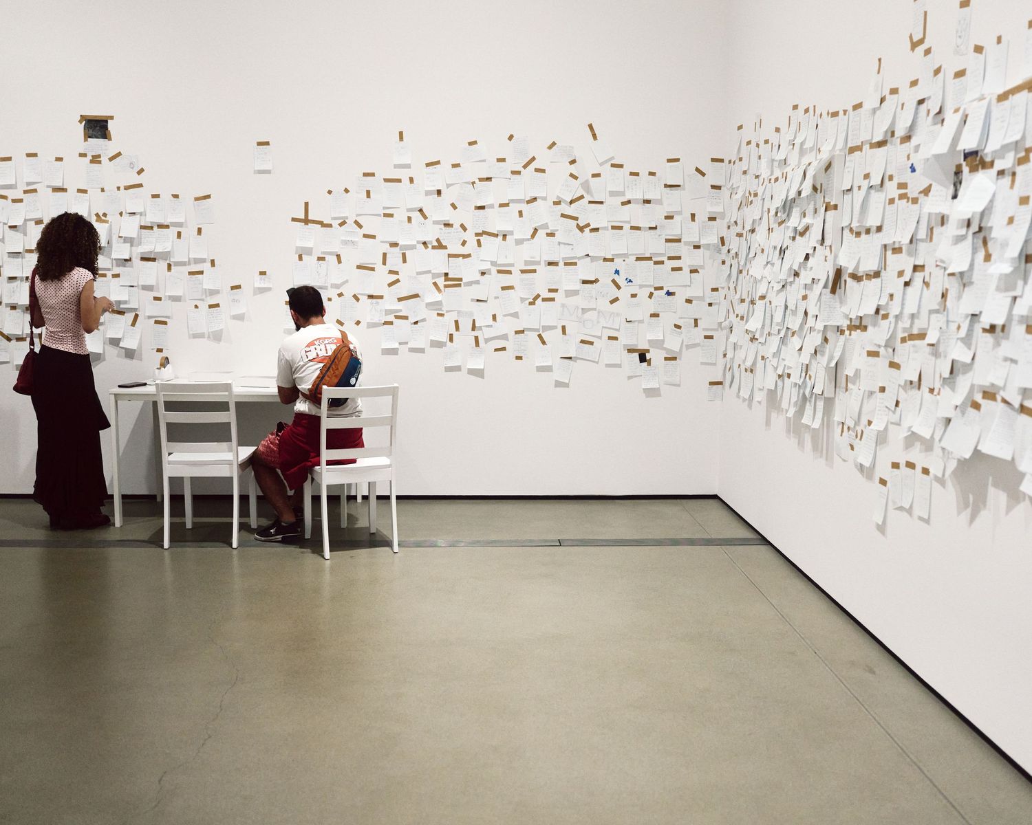 Two visitors write at a desk before a wall of taped notes in Yoko Ono’s My Mommy is Beautiful at The Broad.