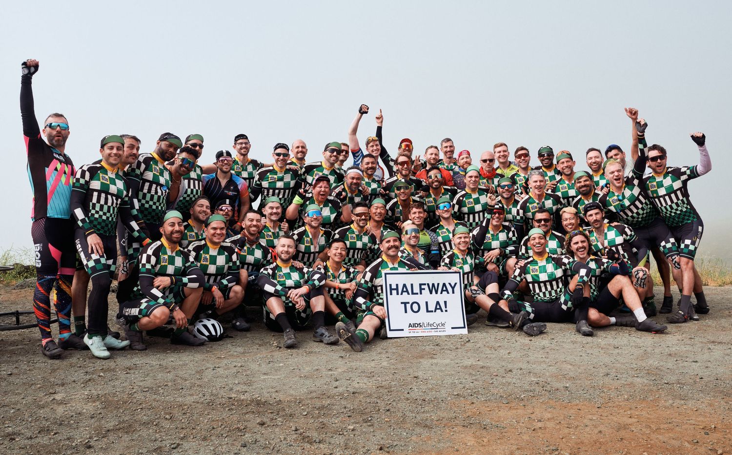 Large group of cyclists in green jerseys gathered for a group photo with 'Halfway to LA' sign.