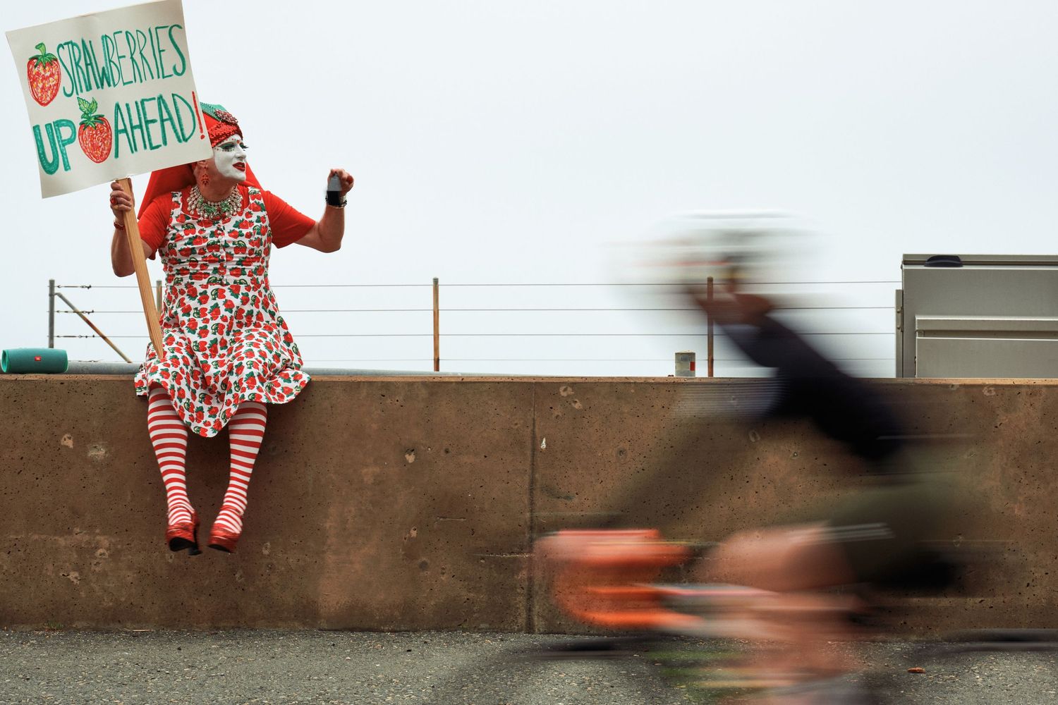 Someone in polka dot dress holds sign while motion-blurred cyclist speeds past concrete wall.