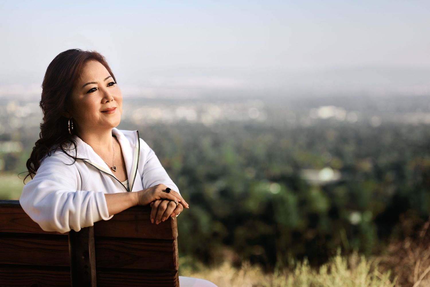 Woman in white shirt sitting on bench with panoramic city view and green hills in background.
