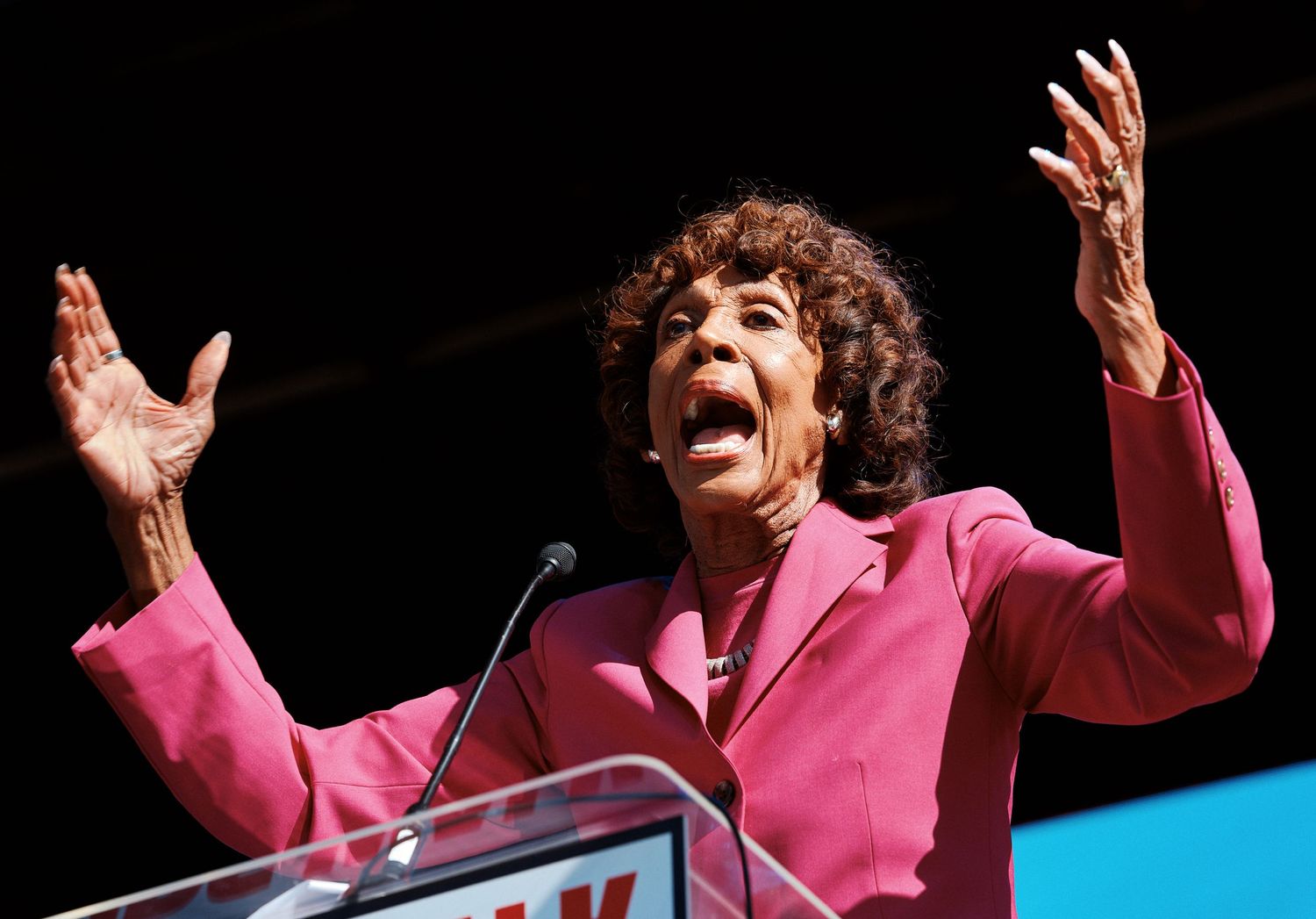 Energetic speaker in bright pink blazer gestures animatedly while delivering a passionate speech at a podium.