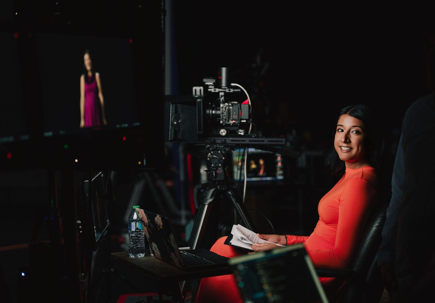 Behind the scenes view of a video production set with camera equipment and a bright red dress in dark lighting.