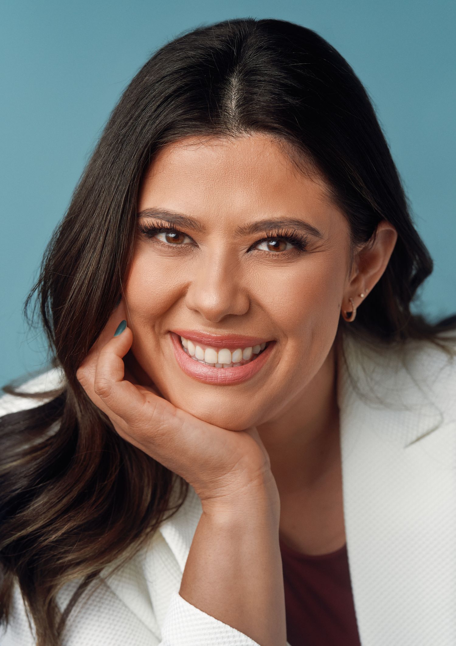 Professional headshot against turquoise background showing warm smile and relaxed pose with hand resting on chin.