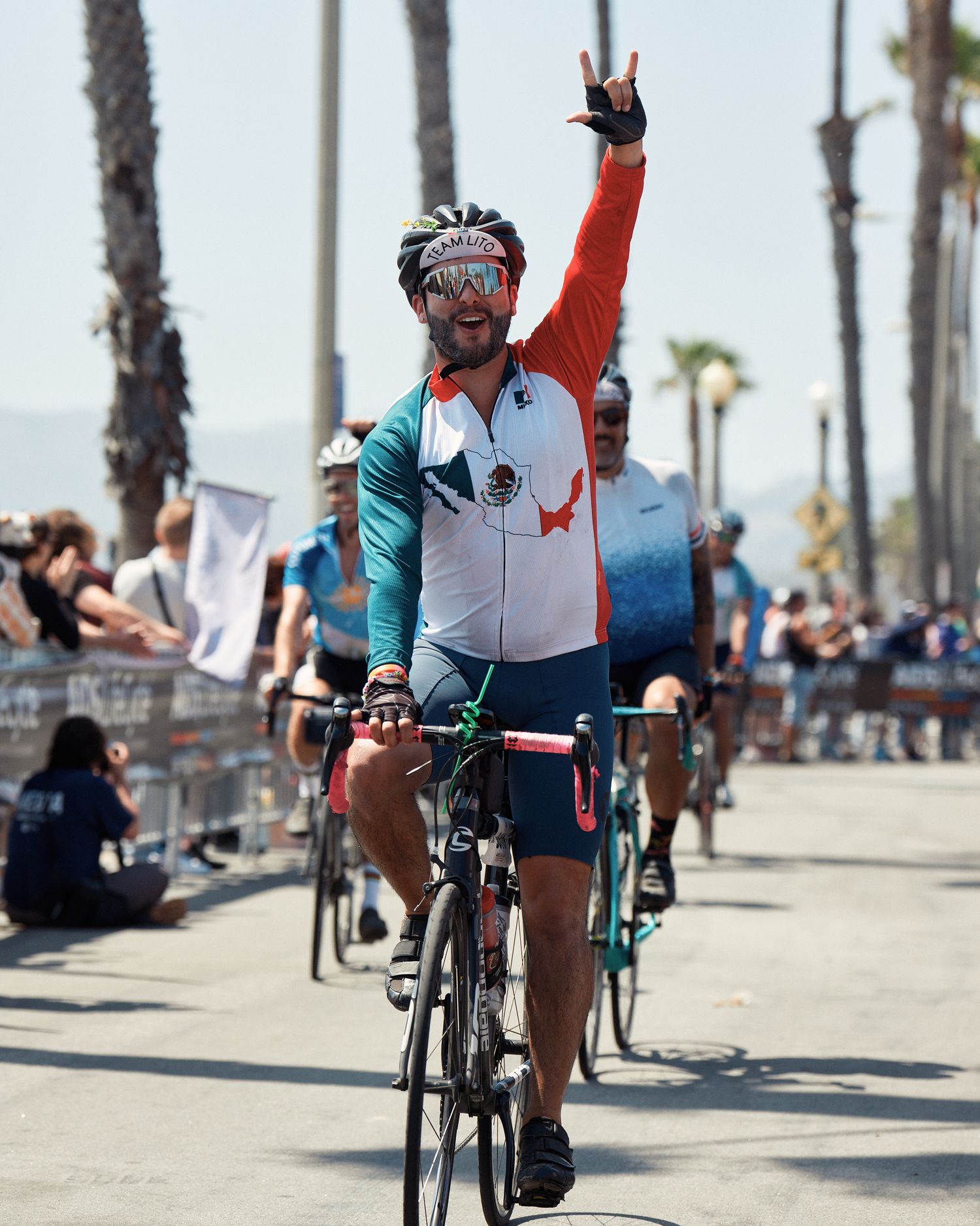 A cyclist in teal and white attire raises arm in victory while riding along a palm tree-lined coastal path.