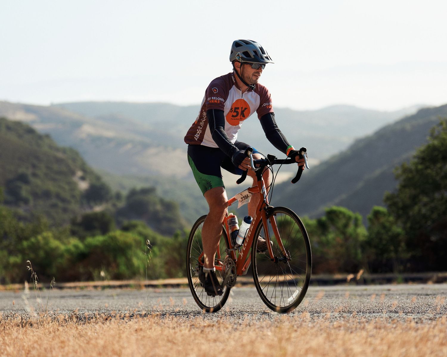 Road cyclist rides orange bicycle on scenic mountain road through rolling hills on sunny California day.