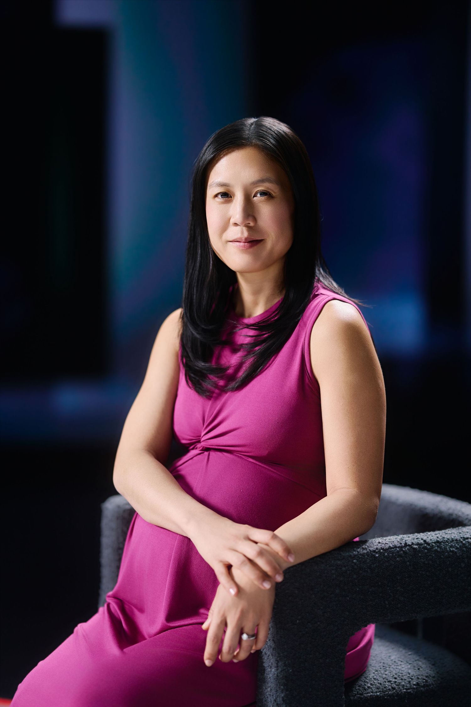 Professional portrait of a person in a magenta sleeveless dress sitting on a dark chair against a blue background.