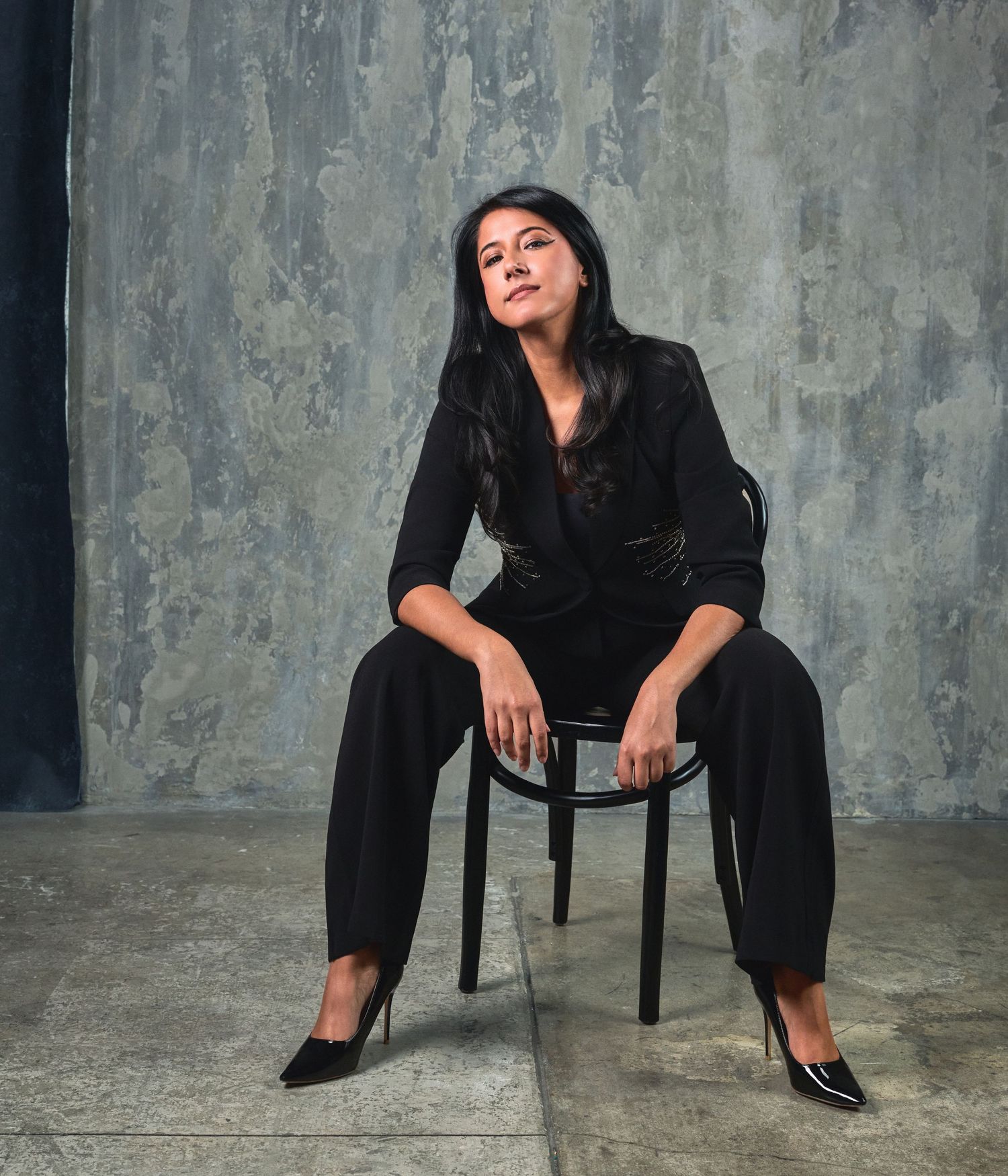 A series of five artistic portrait poses on a chair against a textured gray wall, showcasing elegant black attire.